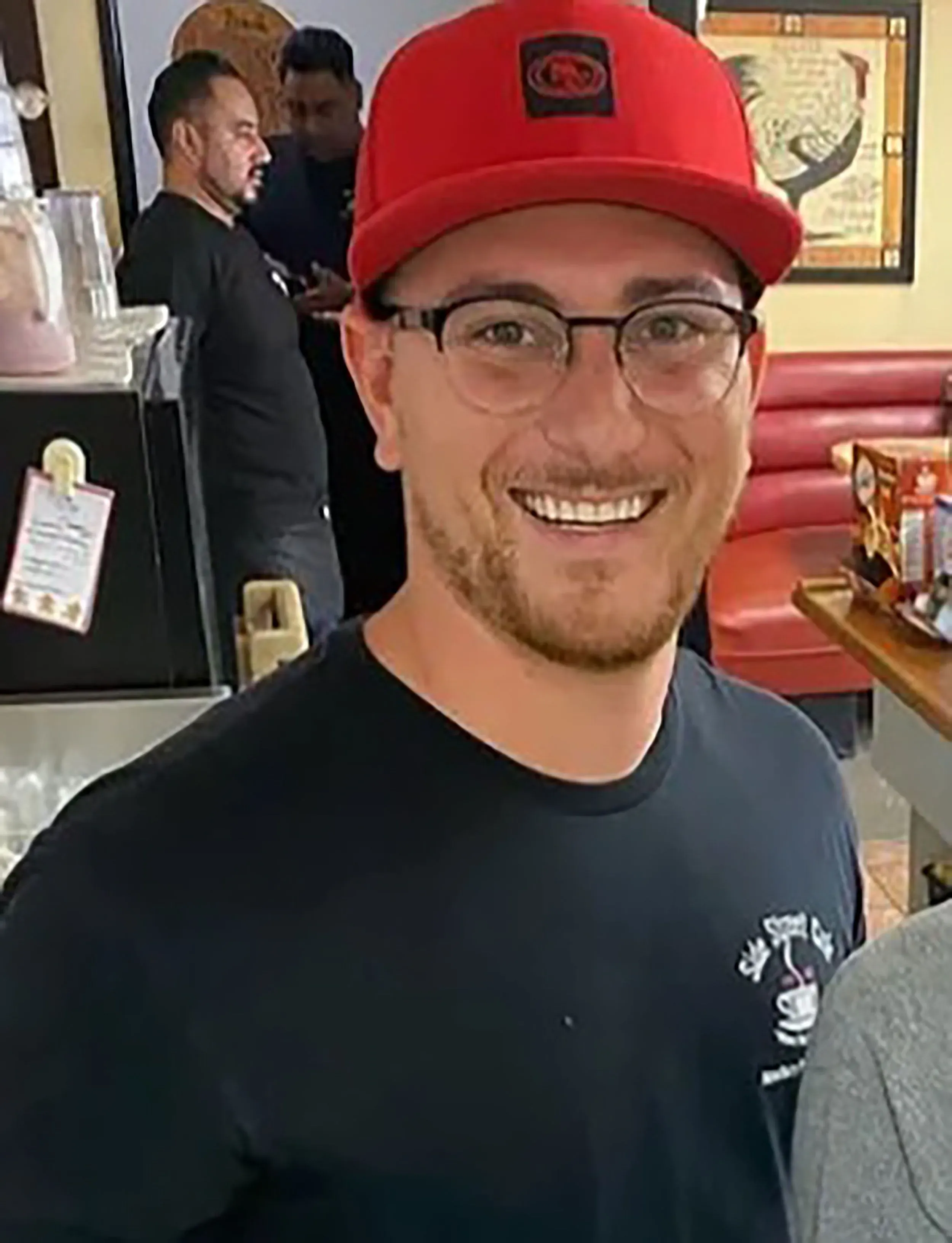 صورة لرجل مبتسم يرتدي قبعة حمراء ونظارات، في بيئة غير رسمية. يمثل جزءًا من مجموعة مرتبطة بحفل توديع العزوبية.