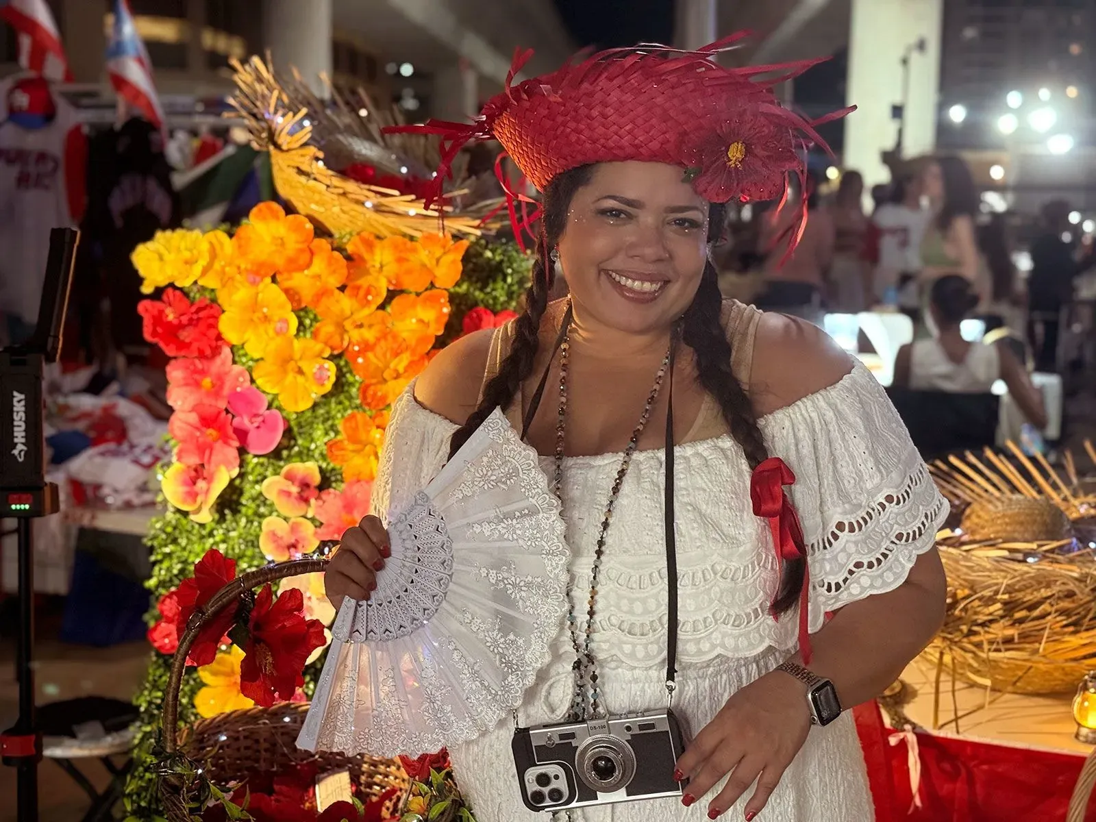 امرأة ترتدي زيًا تقليديًا بورتوريكيًا، تحمل مروحة وتبتسم أمام خلفية مزهرة، تعكس الفخر الثقافي خلال حفل باد باني.