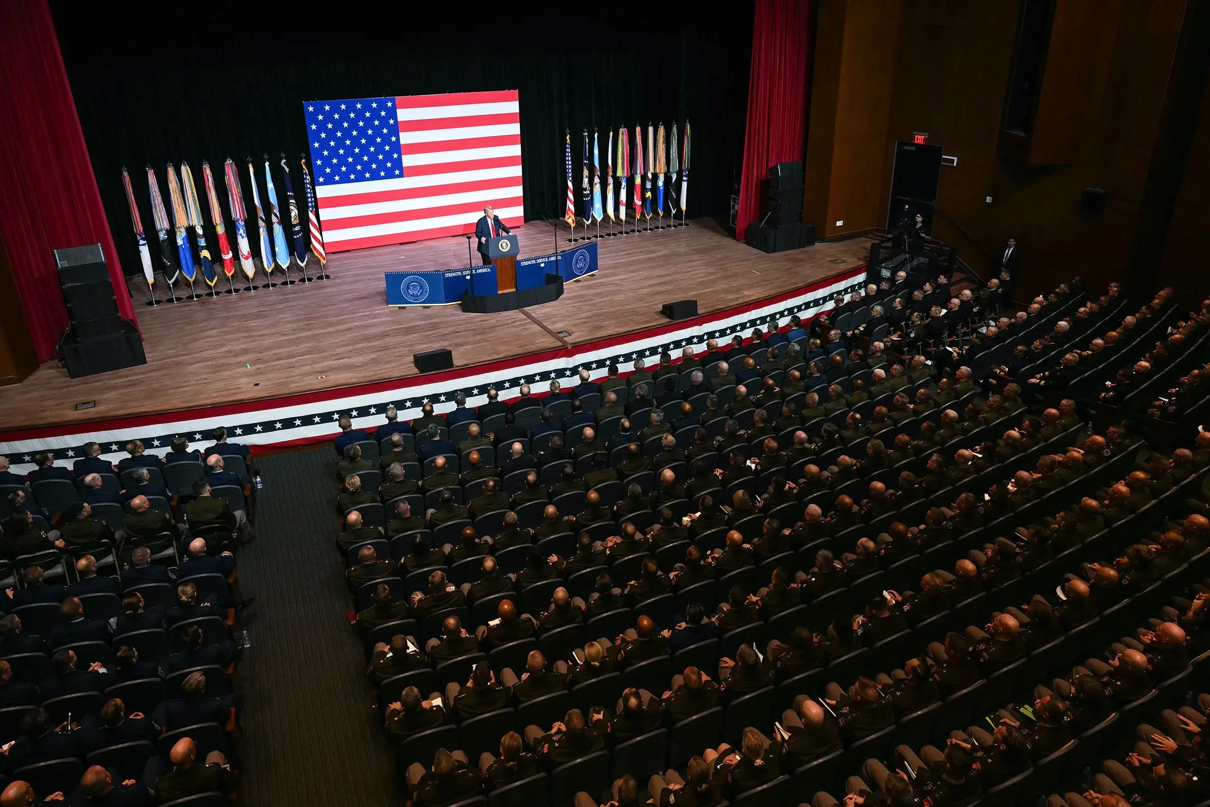 اجتماع عسكري في قاعة كبيرة، حيث يتحدث مسؤول أمام جمهور من الضباط العسكريين، مع العلم الأمريكي خلفه.