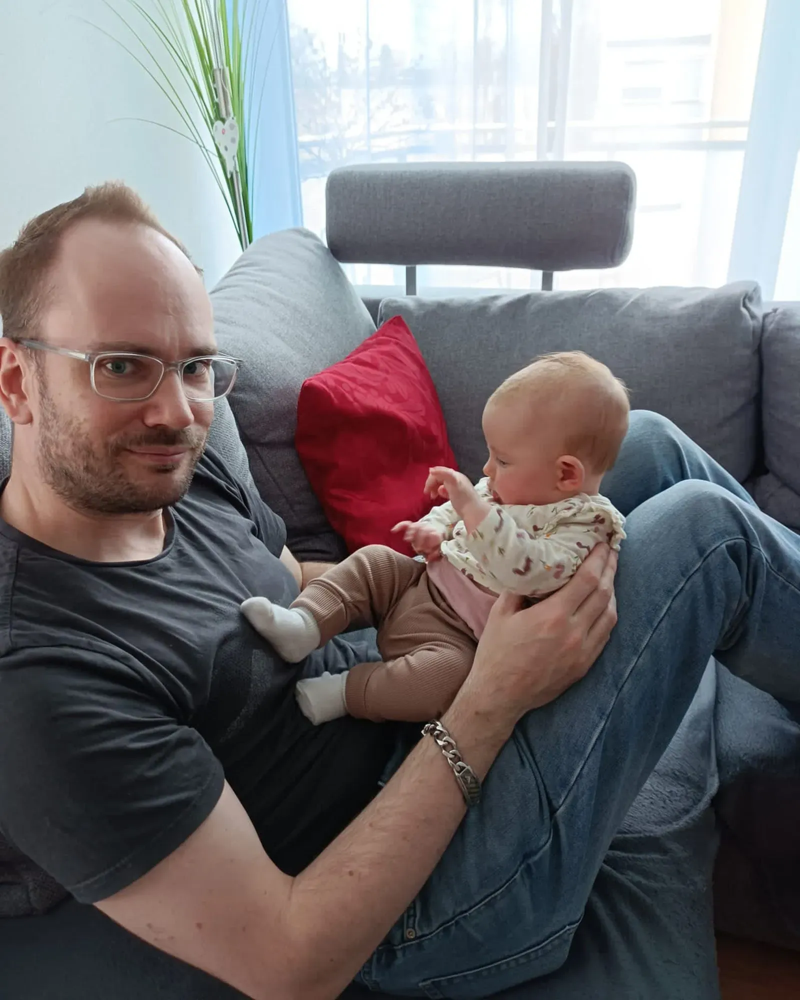 رجل يجلس على أريكة يحمل طفلاً صغيرًا، مع وجود وسادة حمراء خلفه ونوافذ مضاءة في الخلفية. تعكس الصورة لحظة حميمية بين الأب والطفل.