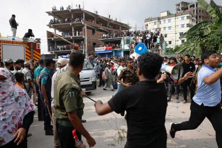 حشود كبيرة من الناس تتجمع في موقع حادث تحطم طائرة تدريب في دكا، مع وجود رجال الشرطة والإسعاف في المكان.