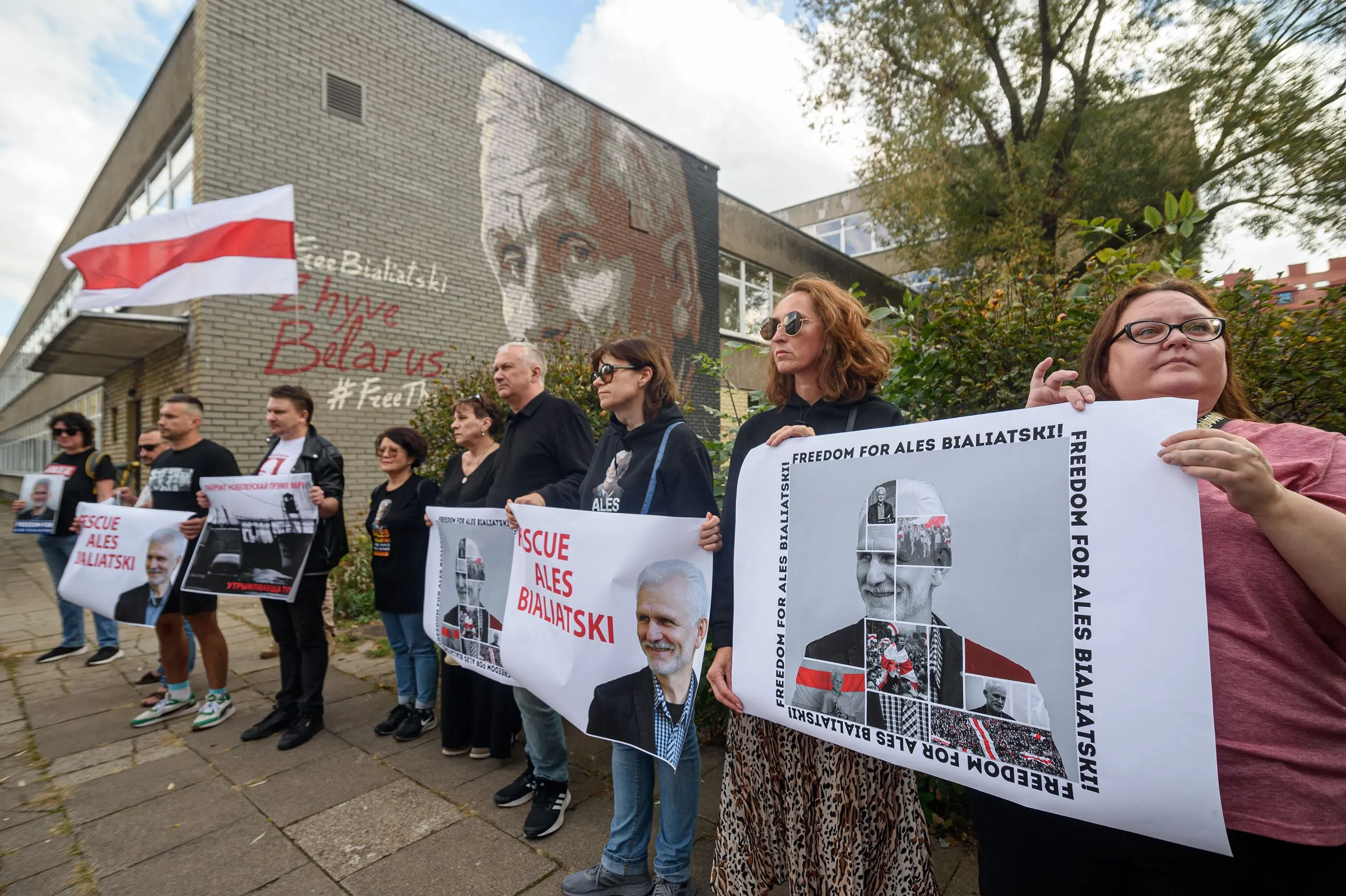 محتجون يحملون لافتات تدعو للإفراج عن أليس بيالياتسكي، مع العلم الوطني البيلاروسي، أمام جدارية له، تعبيرًا عن دعمهم لحقوق الإنسان.