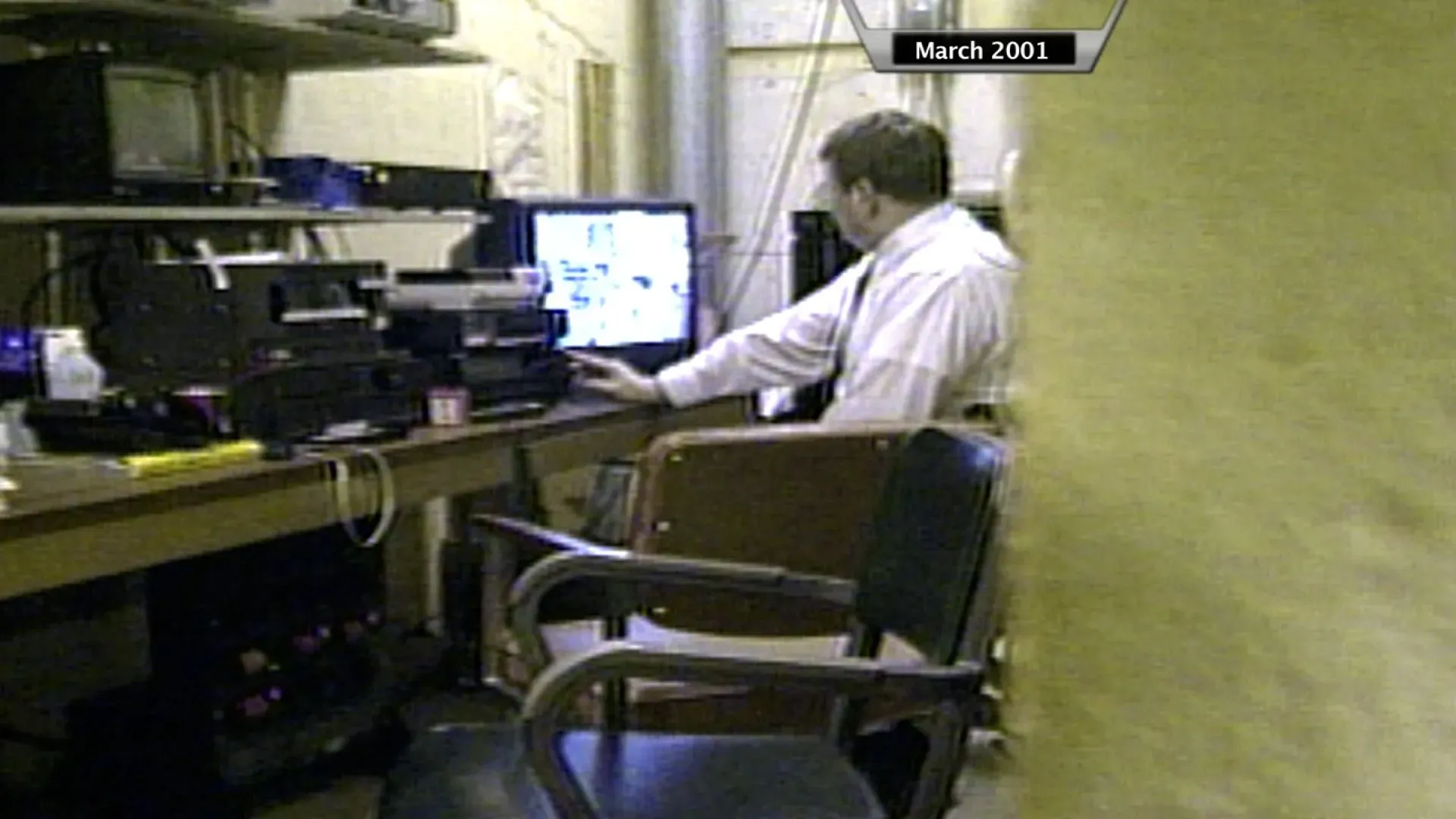 رجل يجلس أمام شاشة كمبيوتر في غرفة تحتوي على معدات تكنولوجية، مع التركيز على العمل الذي يقوم به.