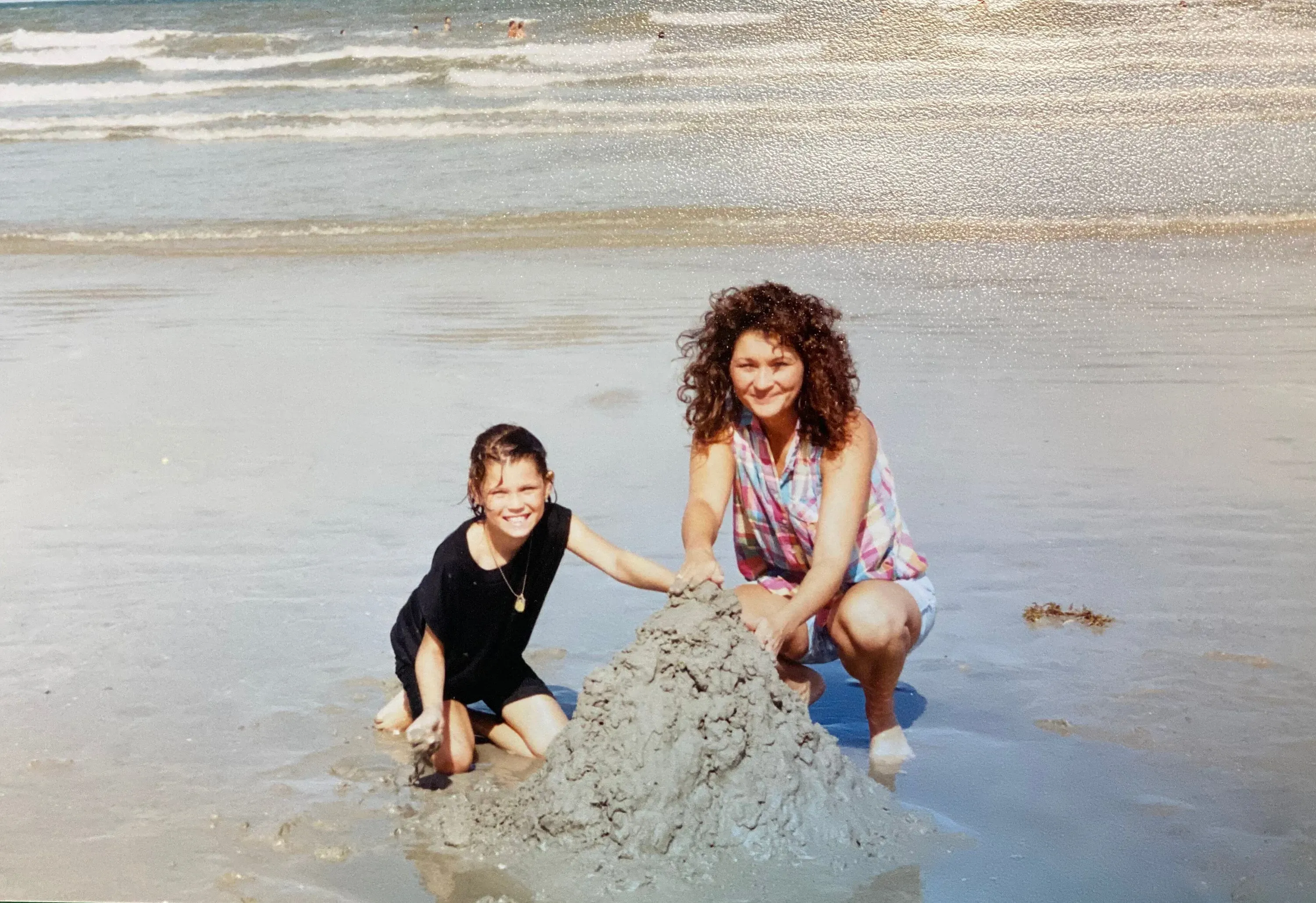 امرأة وفتاة صغيرة تبنيان قلعة رملية على شاطئ البحر، تعبيرات وجههما تعكس السعادة والمرح. الصورة تذكر بذكريات الطفولة والروابط الأسرية.