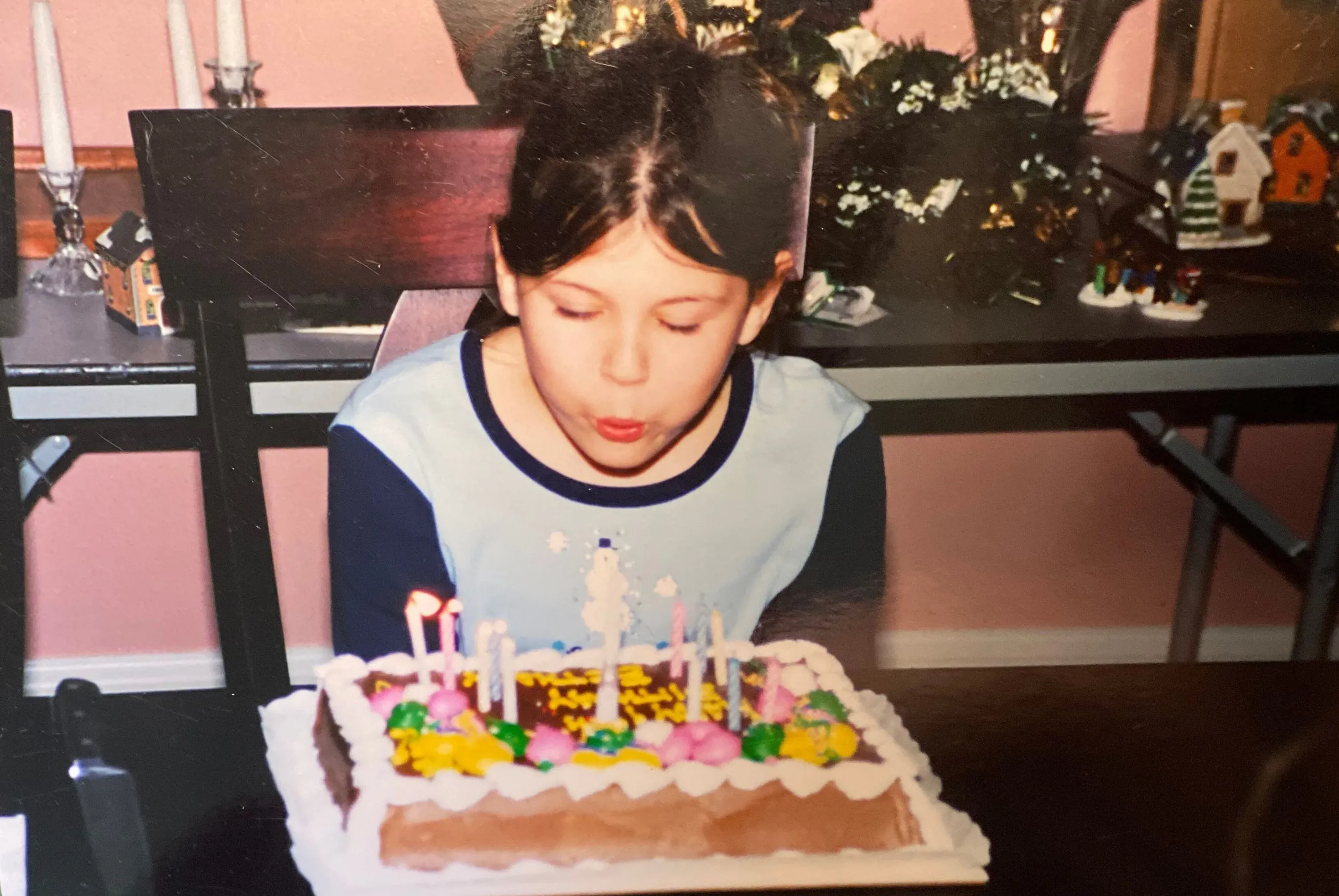 فتاة صغيرة تنفخ شموع عيد ميلادها على كعكة مزينة بألوان زاهية، تعبيرها يدل على الفرح والبراءة، في خلفية منزلية دافئة.