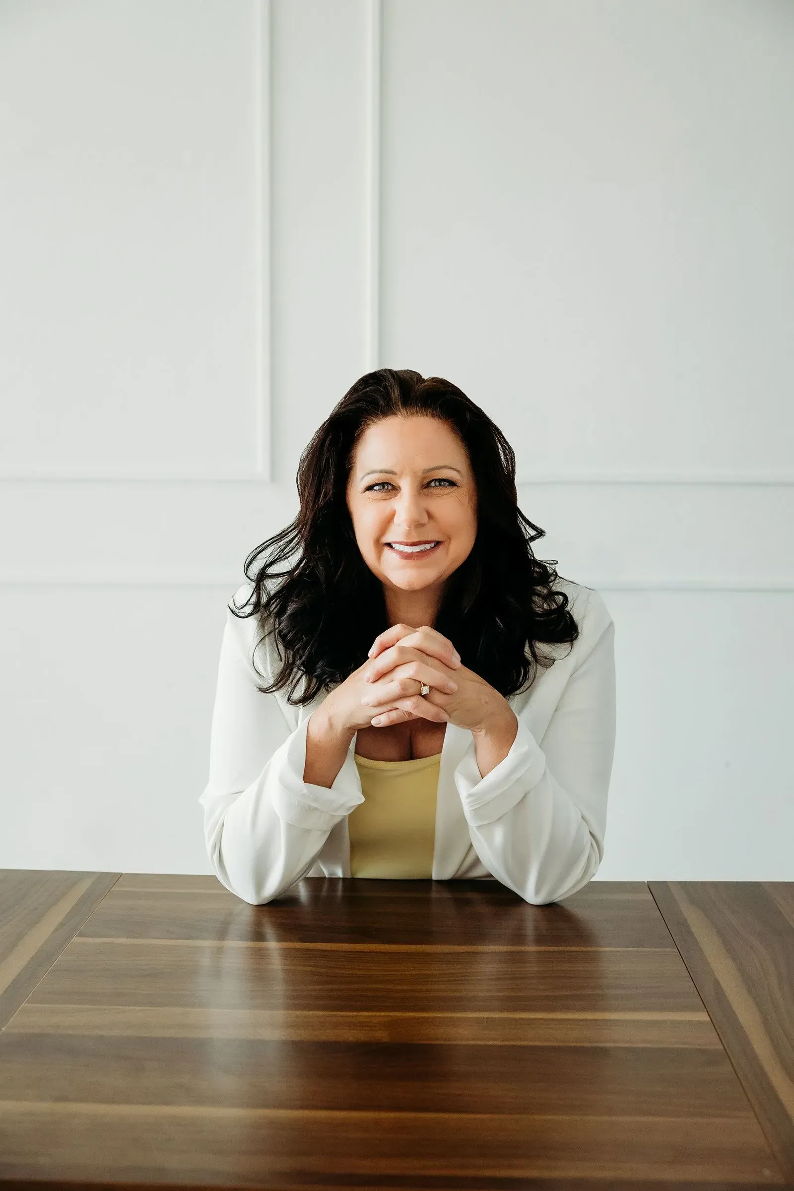 امرأة مبتسمة ترتدي سترة بيضاء، تجلس أمام طاولة خشبية، تعبر عن الثقة والإيجابية، تتحدث عن أهمية التغييرات الصغيرة لتحقيق الأهداف.