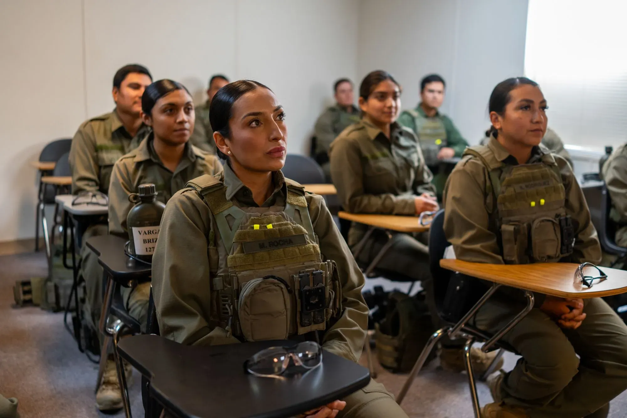 مجموعة من المتدربين في أكاديمية حرس الحدود الأمريكية، يرتدون زيًا رسميًا، يستمعون إلى محاضرة في قاعة دراسية.