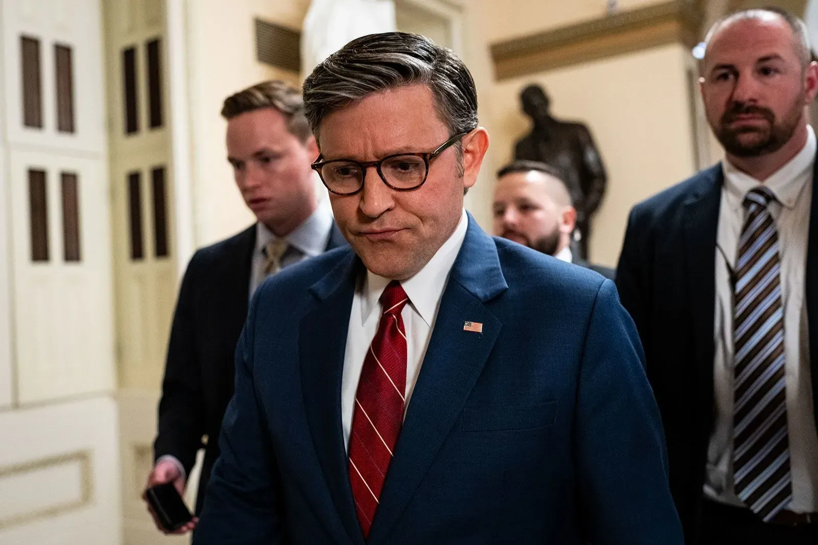 رجل يرتدي بدلة زرقاء ونظارات، يبدو عليه القلق، يسير في ممر مع مجموعة من الأشخاص، وسط أجواء سياسية مشحونة.