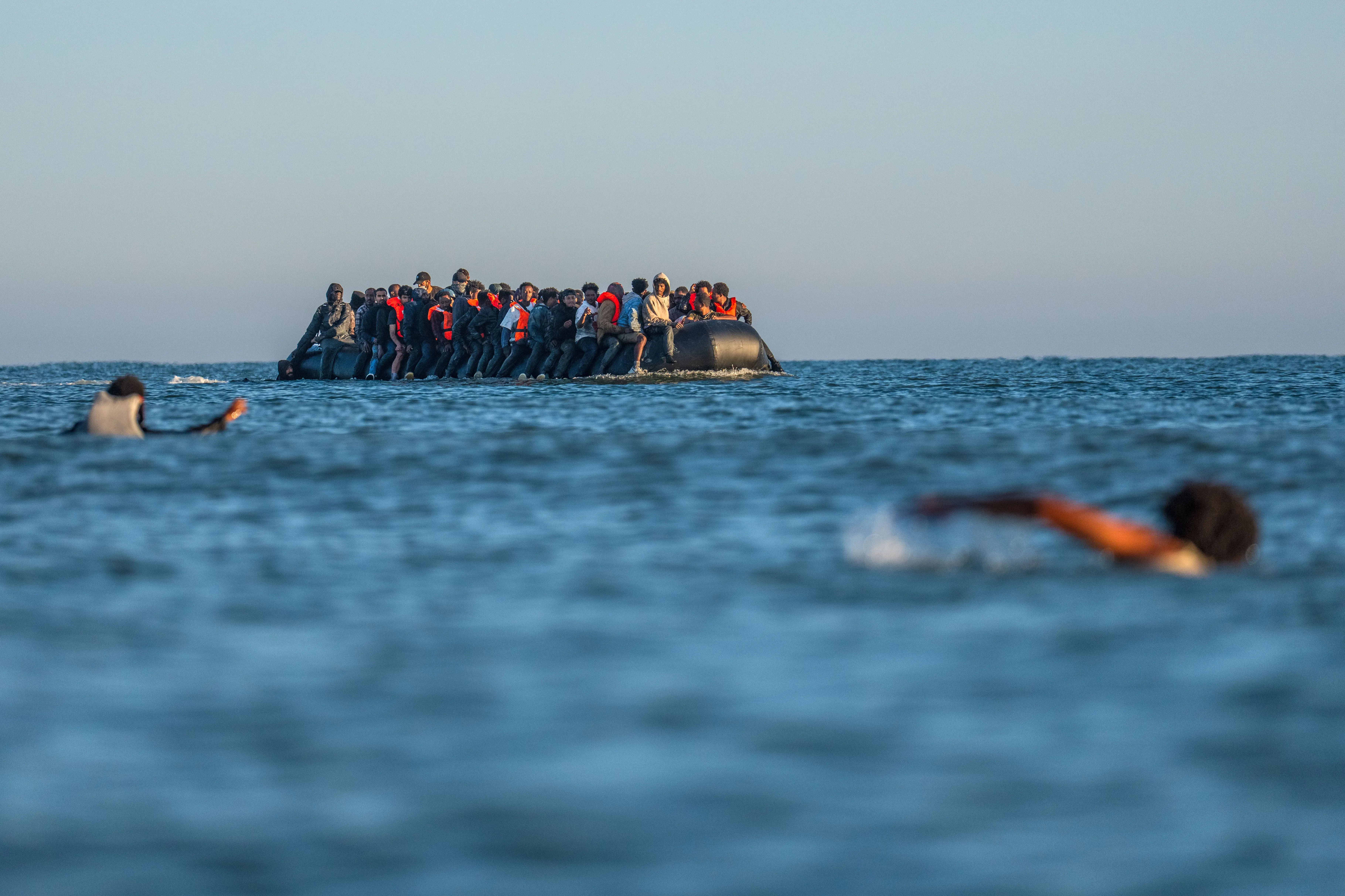مجموعة من اللاجئين في قارب مطاطي صغير يعبرون البحر، مع شخص آخر يسبح بالقرب منهم، في سياق أزمة الهجرة في بريطانيا.