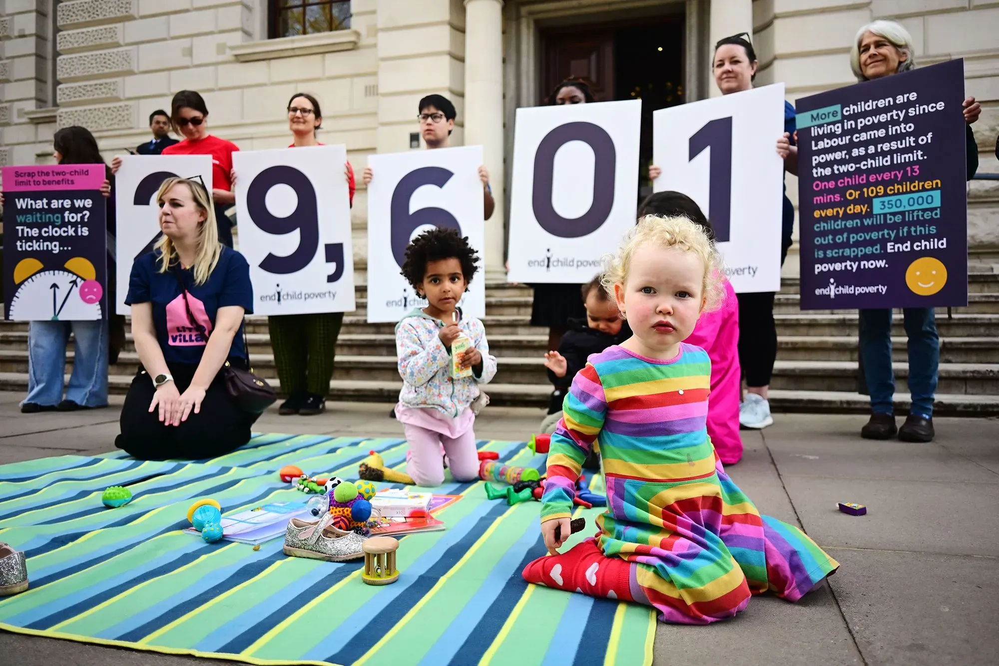 مجموعة من المحتجين أمام مبنى، يحملون لافتات تشير إلى أرقام الفقر بين الأطفال في المملكة المتحدة، مع وجود طفل صغير يلعب على سجادة.