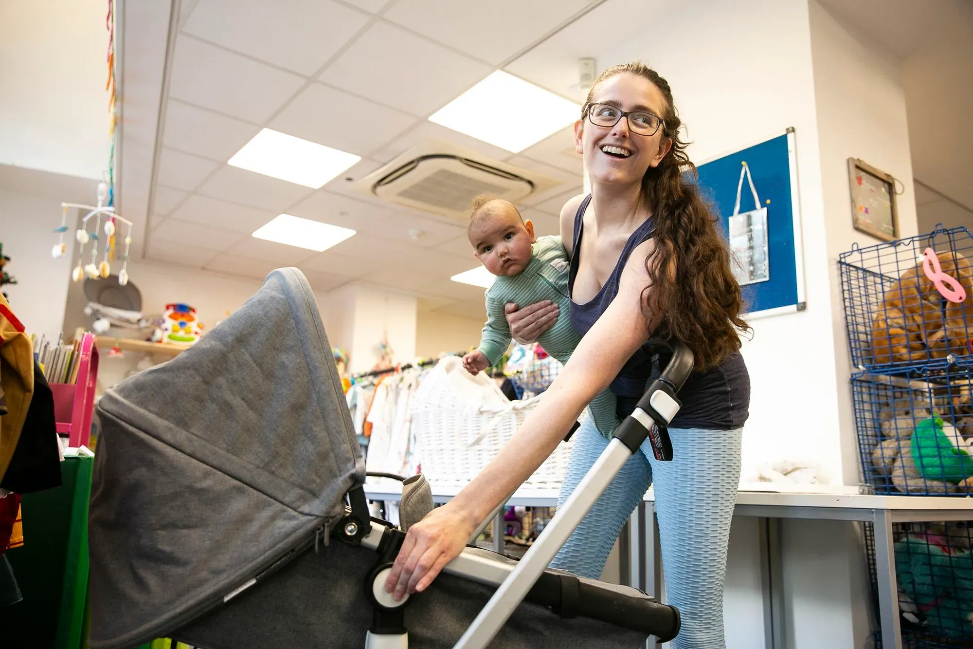 امرأة شابة تحمل طفلًا في حضنها وتبتسم بينما تدفع عربة أطفال في مركز مجتمعي، تعكس تحديات الأمهات العازبات في مواجهة الفقر.