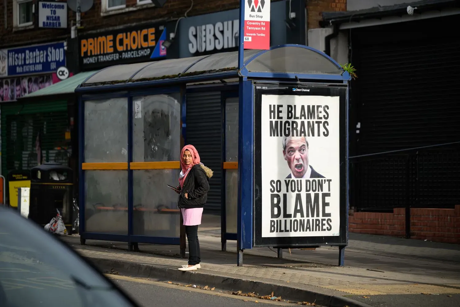 امرأة ترتدي حجابًا تقف في محطة حافلات بجانب لافتة تتهم نايجل فاراج بإلقاء اللوم على المهاجرين لتجنب لوم المليارديرات.