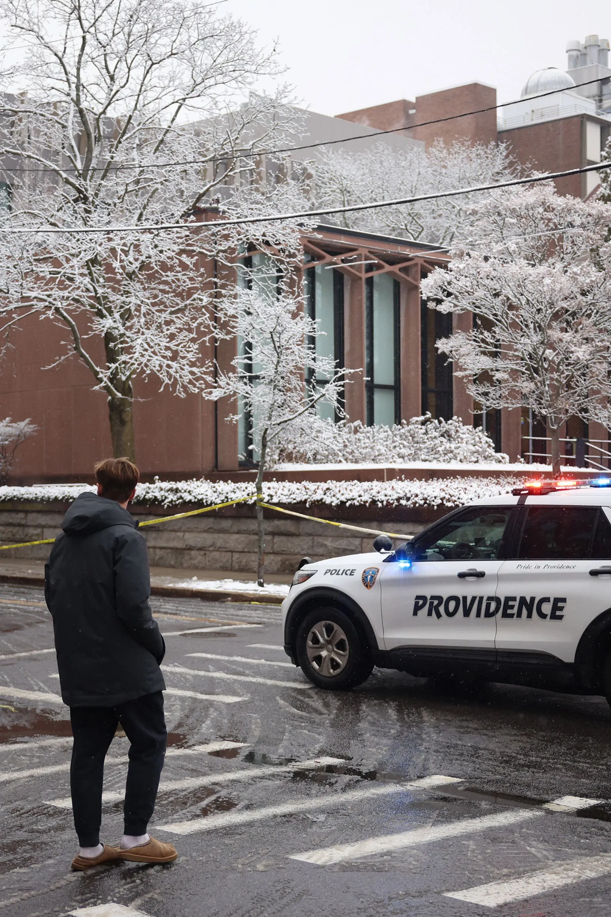 طالب يقف أمام سيارة شرطة في جامعة براون، محاطًا بأشجار مغطاة بالثلوج، مع وجود شريط تحذيري يشير إلى موقع حادث إطلاق نار سابق.