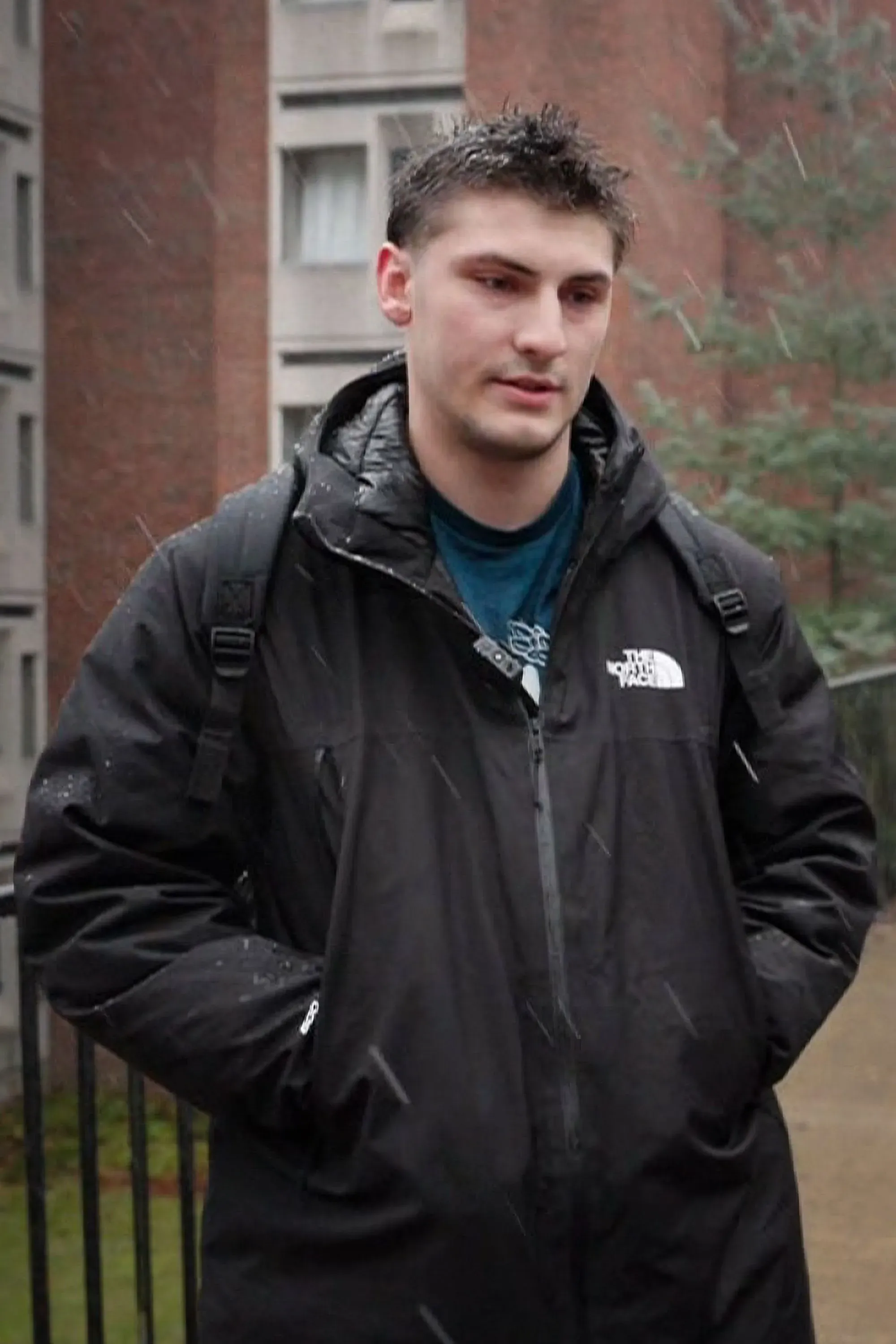 طالب في جامعة براون يقف في الحرم الجامعي تحت المطر، معبرًا عن مشاعر القلق بعد حادث إطلاق النار الأخير.