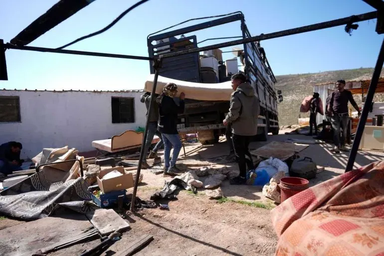 مجموعة من الأشخاص يقومون بنقل الأثاث من منزلهم في منطقة تعرضت للمصادرة في الضفة الغربية، تعبيرًا عن معاناتهم بسبب التوسعات الاستيطانية.