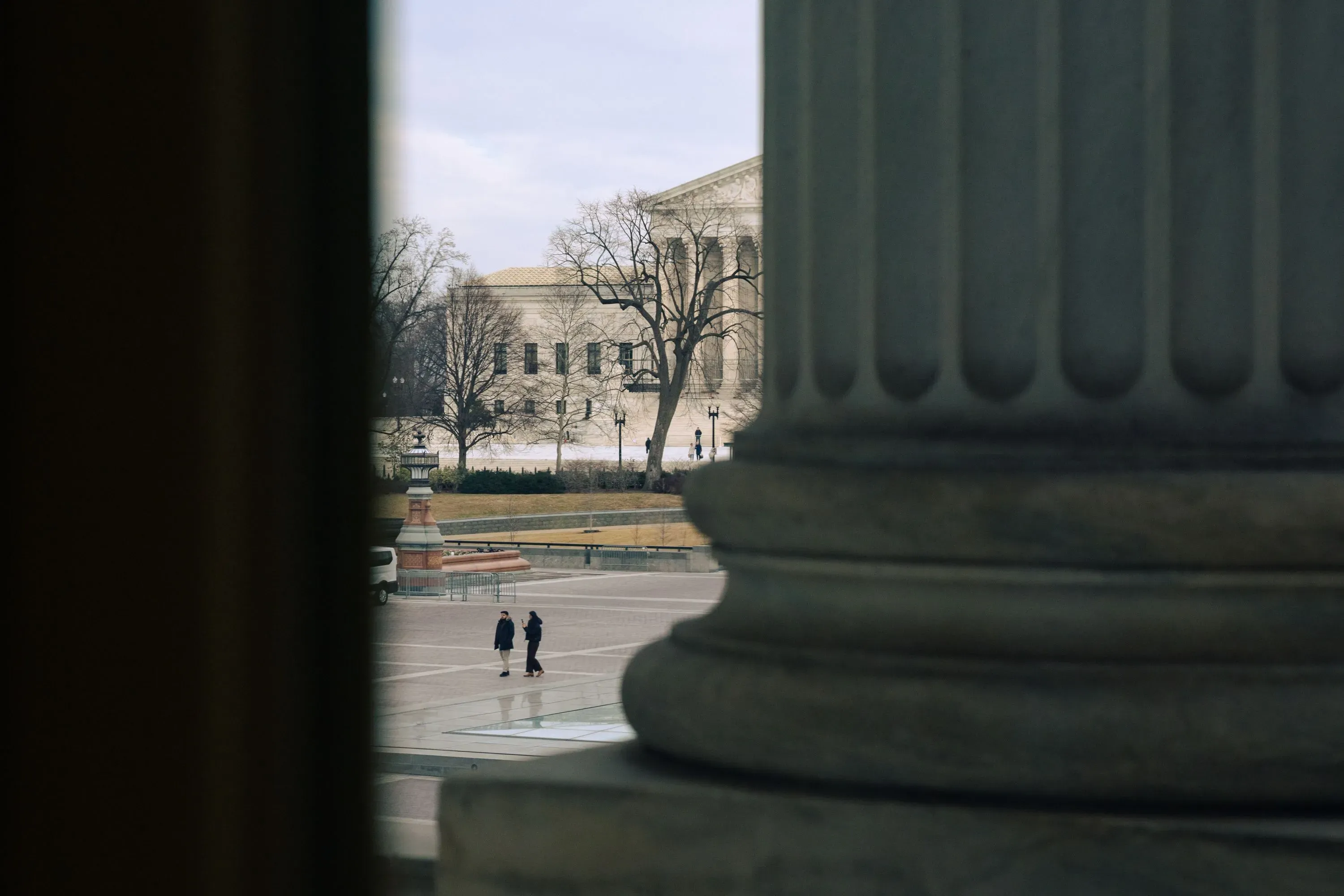 مشهد لشارع أمام مبنى المحكمة العليا، حيث يسير شخصان تحت الأشجار، مع أعمدة معمارية بارزة في المقدمة.