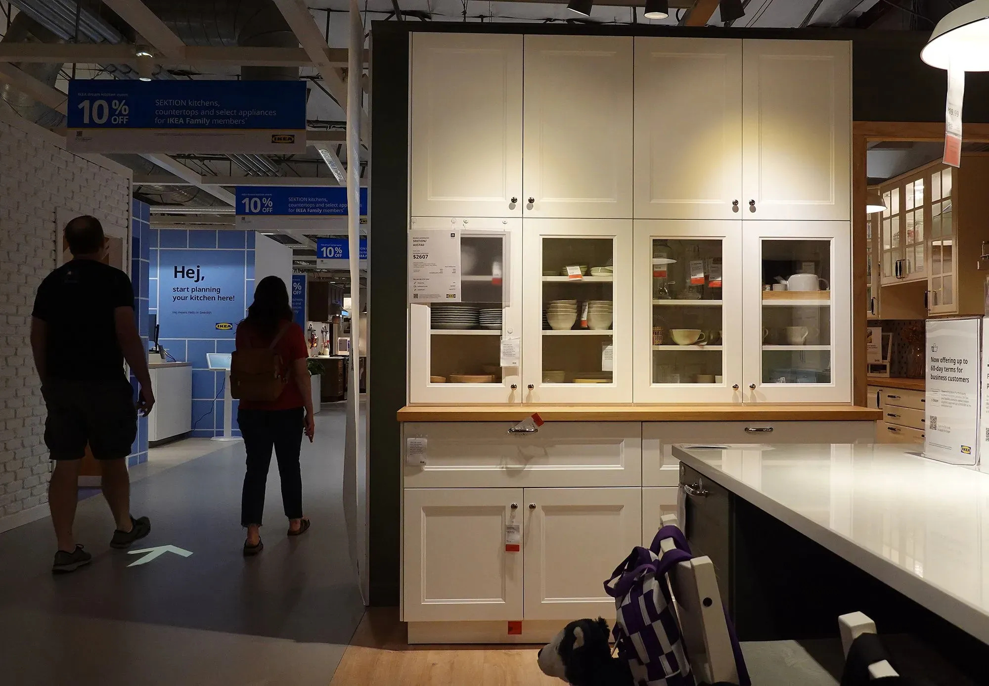 صورة داخل متجر أثاث تعرض خزائن مطبخ بيضاء مع أرفف زجاجية، بينما يتجول زبائن في الممرات. تشير اللافتات إلى خصم 10% على المنتجات.
