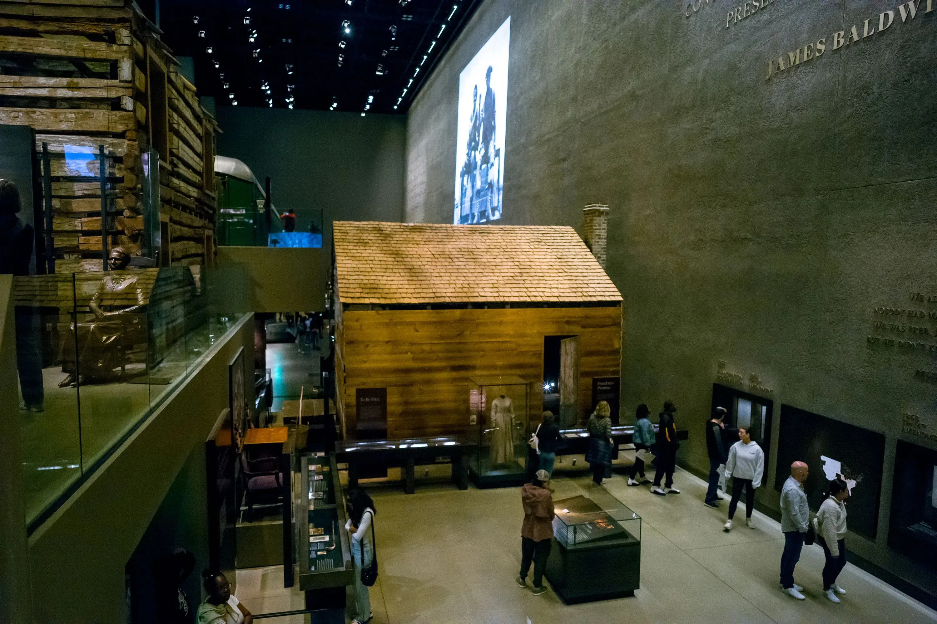 معرض في متحف يبرز تاريخ العبودية في أمريكا، يظهر بيت خشبي قديم وأشخاص يتجولون بين المعروضات التعليمية.