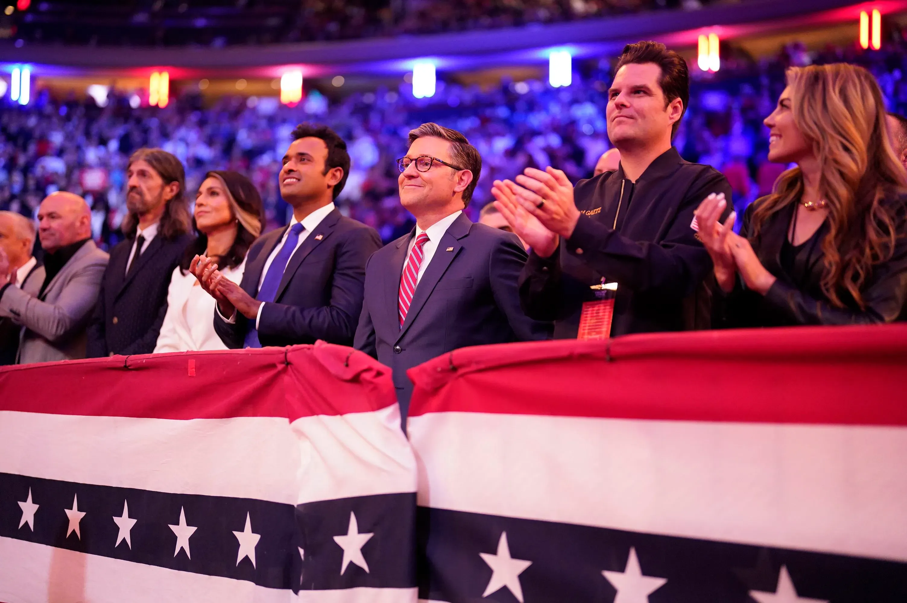 مجموعة من الأشخاص في حدث سياسي، يصفقون ويظهرون تفاعلهم مع الأجواء، بينما تتواجد خلفهم لافتة تحمل نجومًا وخطوطًا ملونة.