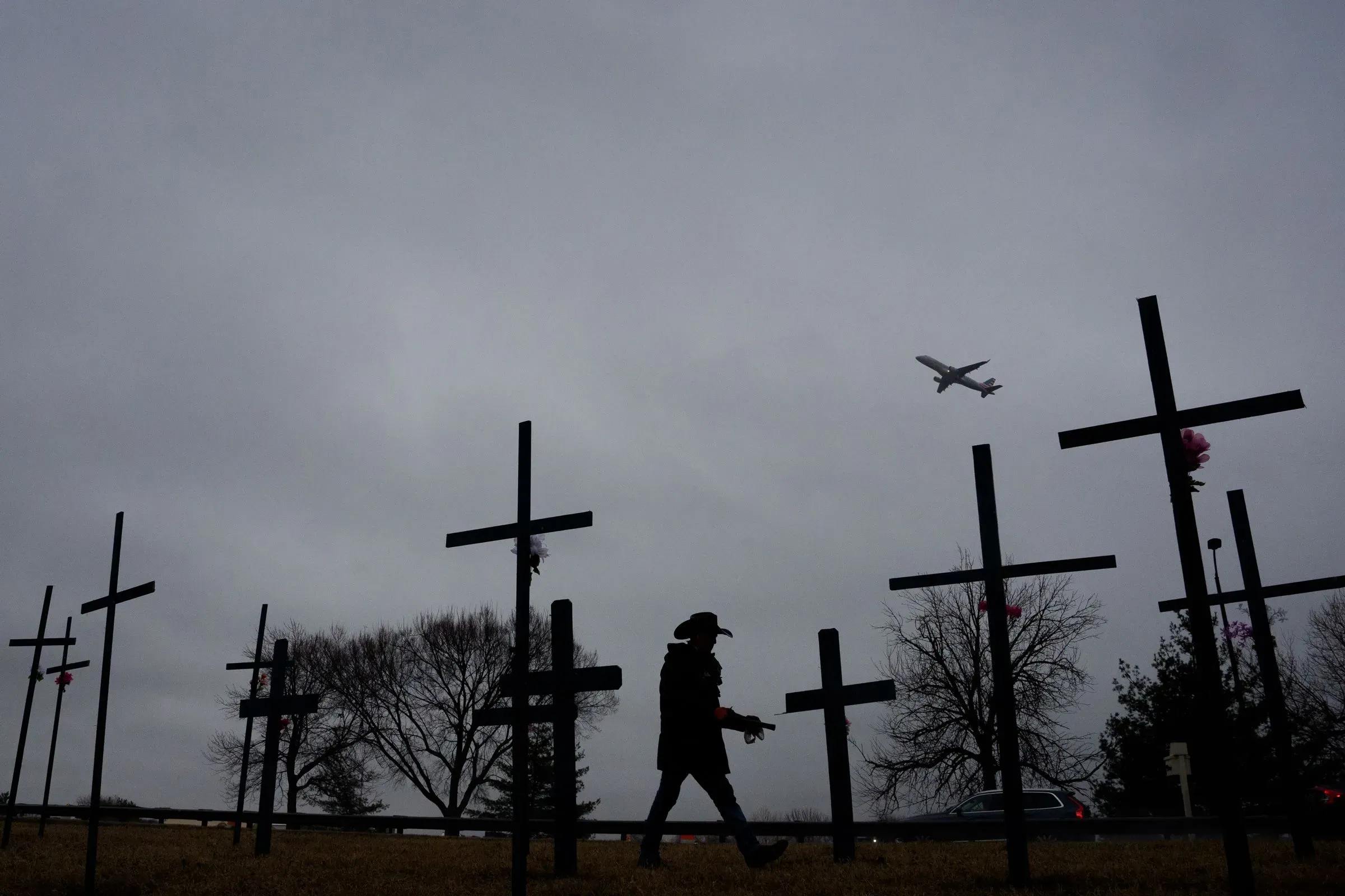 شخص يسير بجوار صلبان تذكارية في يوم غائم، بينما تحلق طائرة فوقهم، مما يرمز إلى الحوادث الجوية وتأثيرها على السلامة.