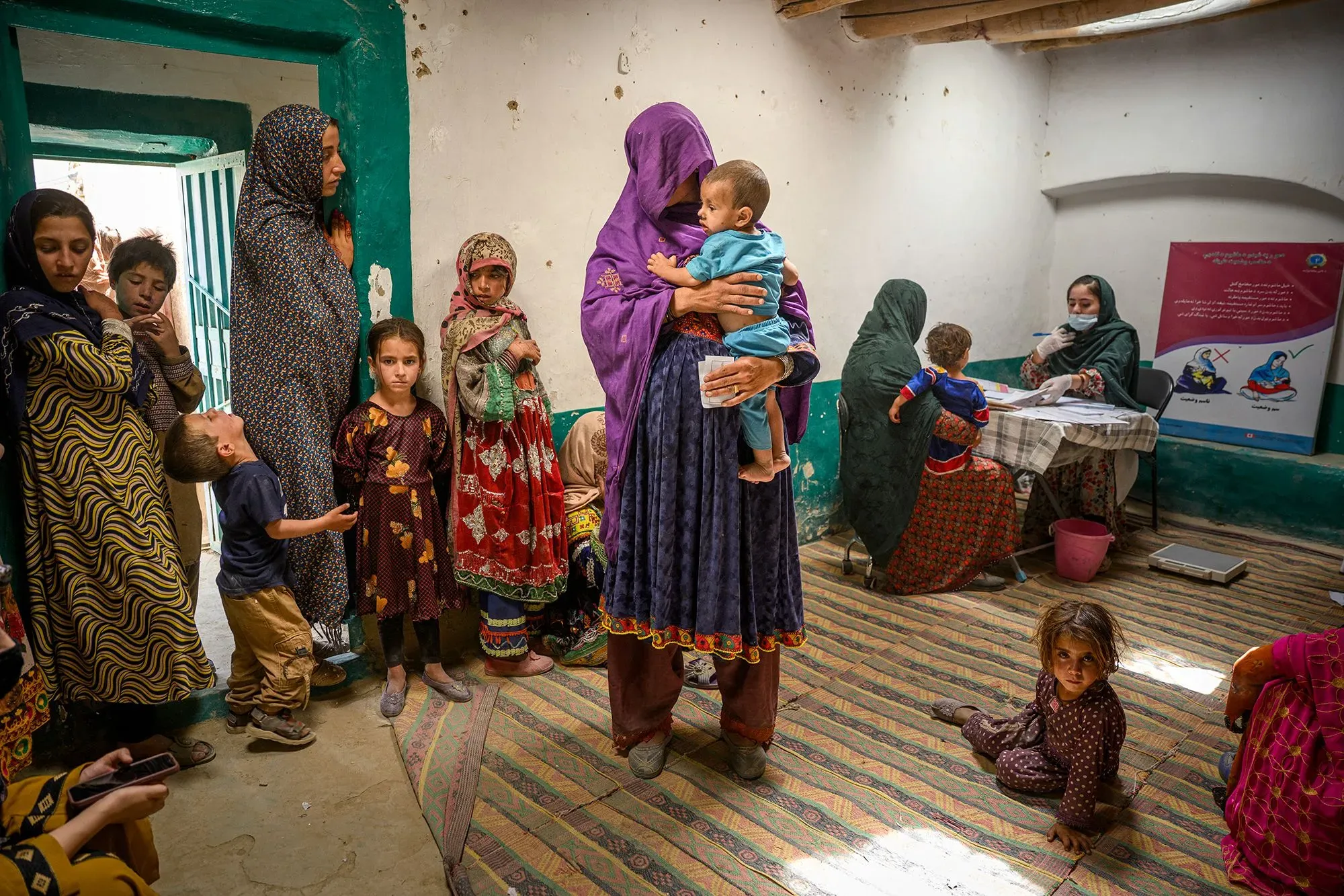 امرأة تحمل طفلاً في مركز صحي، بينما تنتظر مجموعة من النساء والأطفال للحصول على الرعاية الصحية والتطعيمات اللازمة.