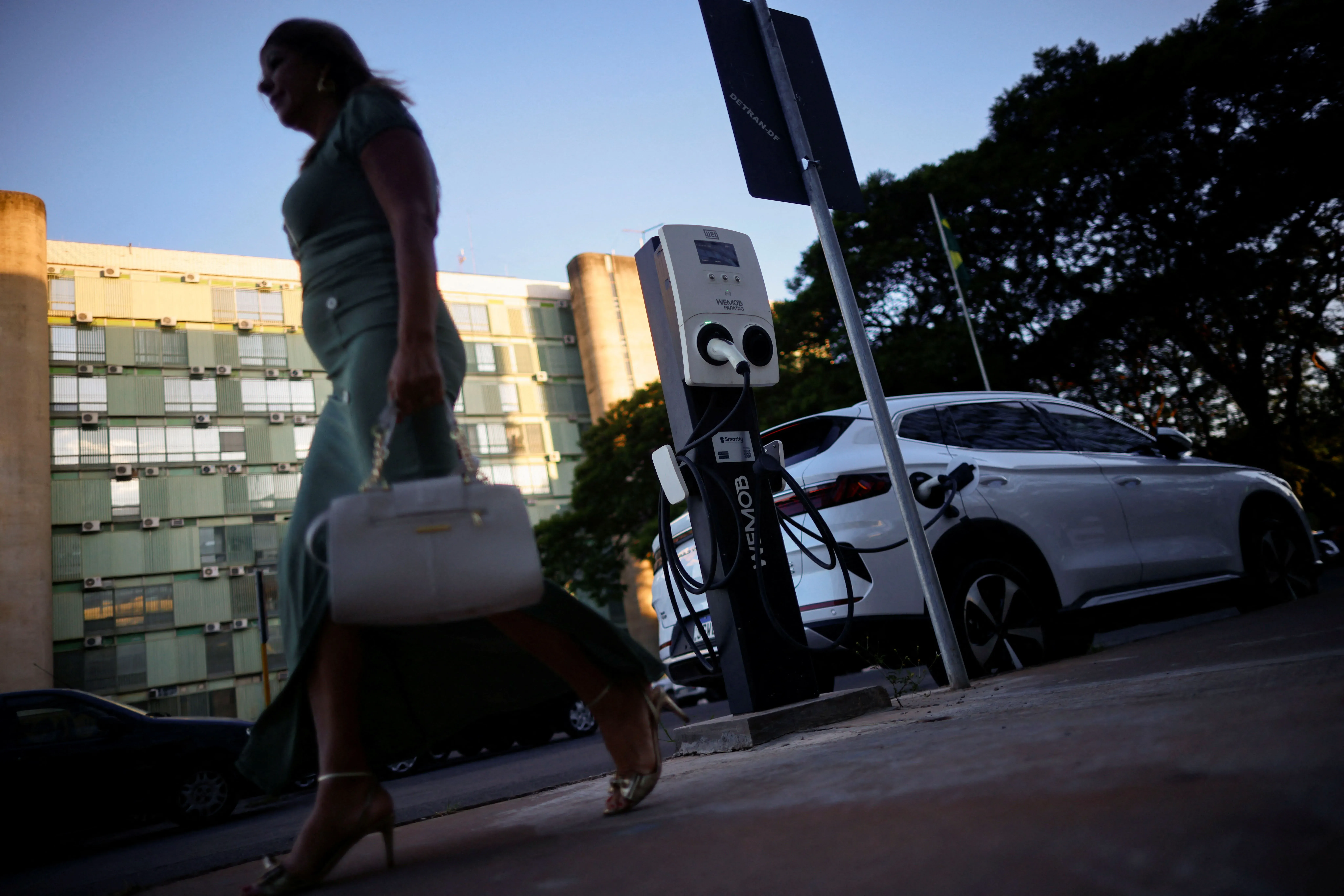 امرأة تسير بجانب محطة شحن للسيارات الكهربائية، مع سيارة بيضاء متوقفة بجانبها، في بيئة حضرية تعكس التطور التكنولوجي.