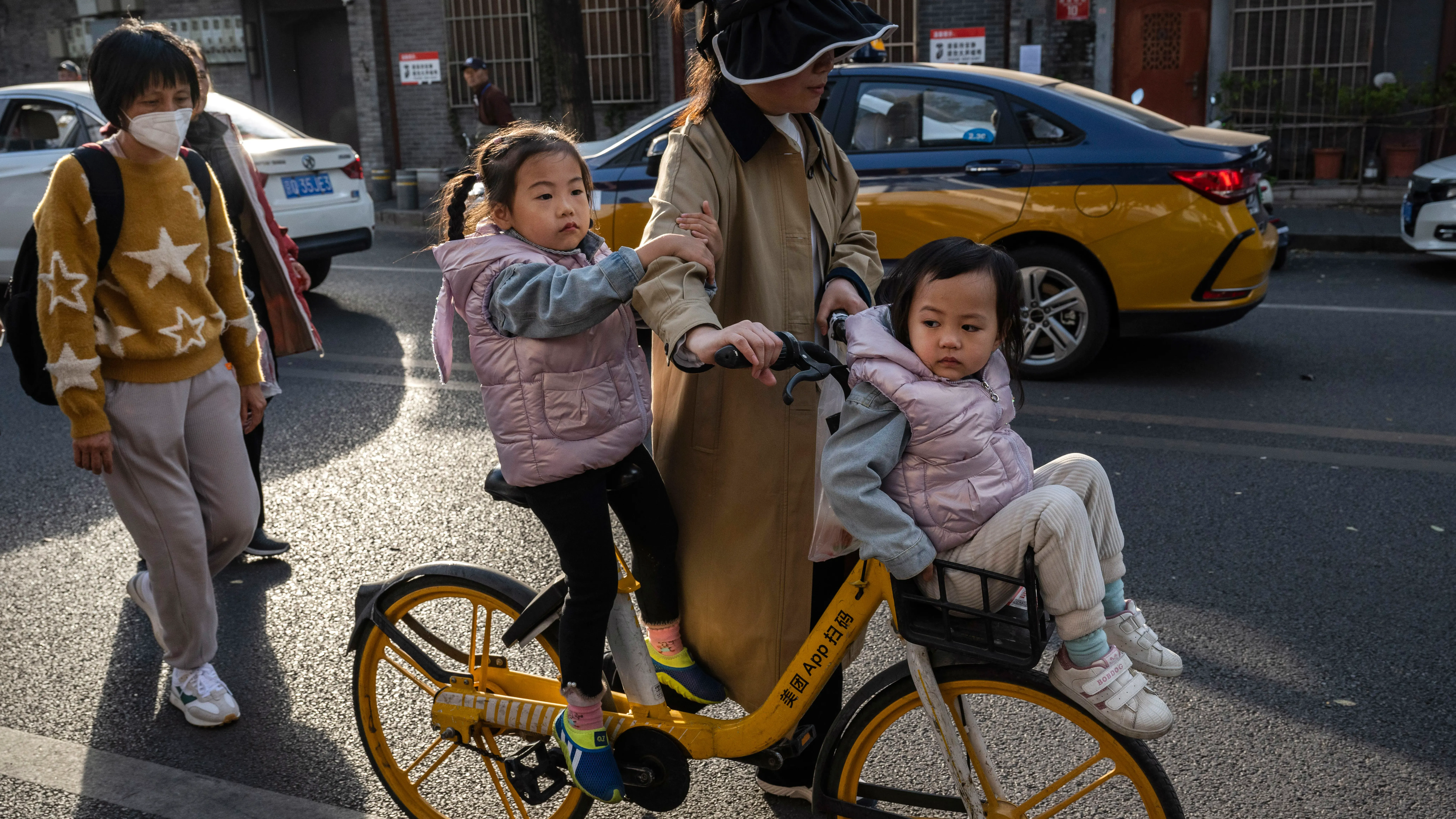 امرأة تقود دراجة مع طفلين يرتديان سترات وردية في شارع مزدحم بالصين، مع سيارات وأشخاص آخرين في الخلفية.