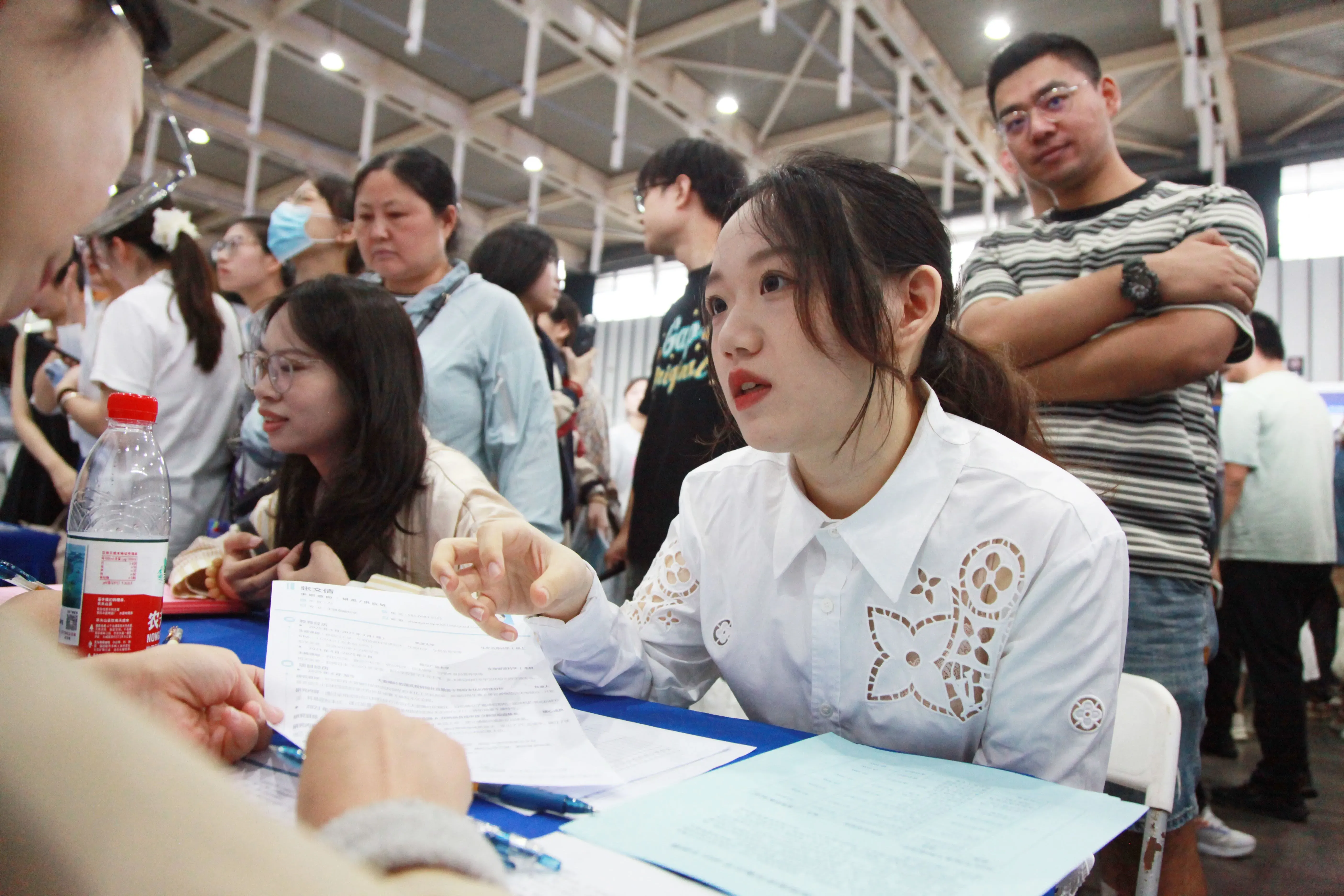 امرأة شابة ترتدي قميصًا أبيض وتجلس أمام طاولة، تتحدث مع شخص آخر في حدث توظيف مزدحم، مع وجود حشود من الناس في الخلفية.