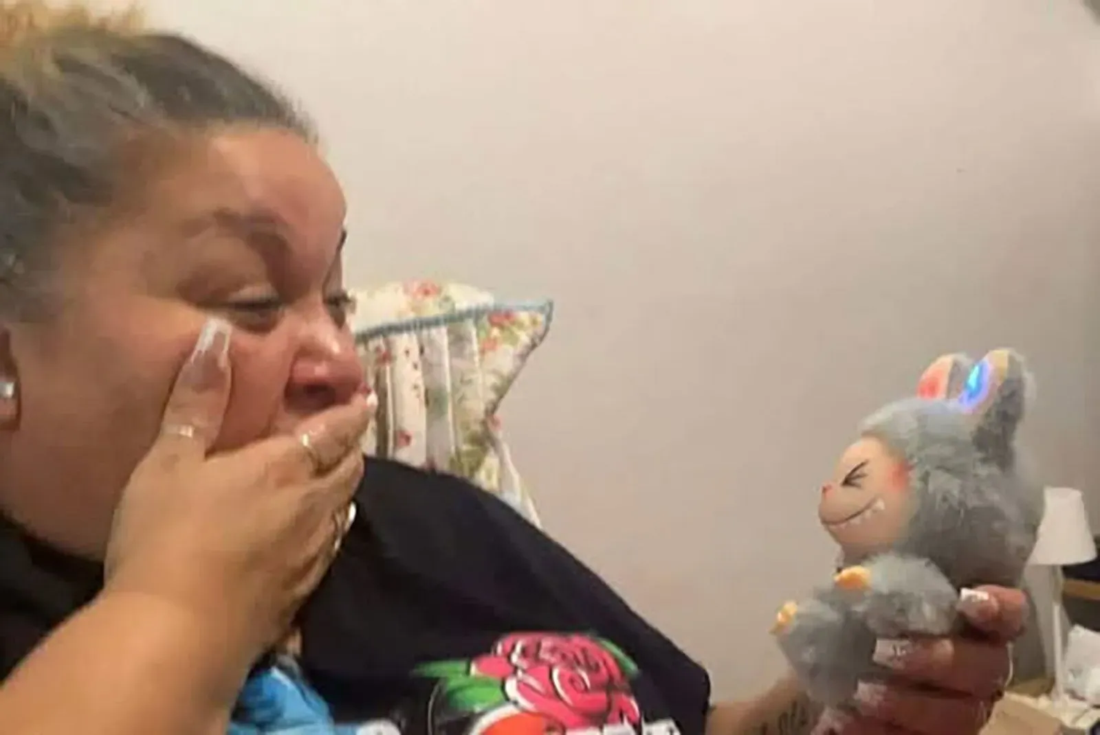 امرأة تعبر عن دهشتها وسعادتها أثناء احتضان دمية "لافوفو" ذات الأذنين المضيئتين، مما يعكس جاذبيتها الفريدة والسحر القبيح للدمى المقلدة.