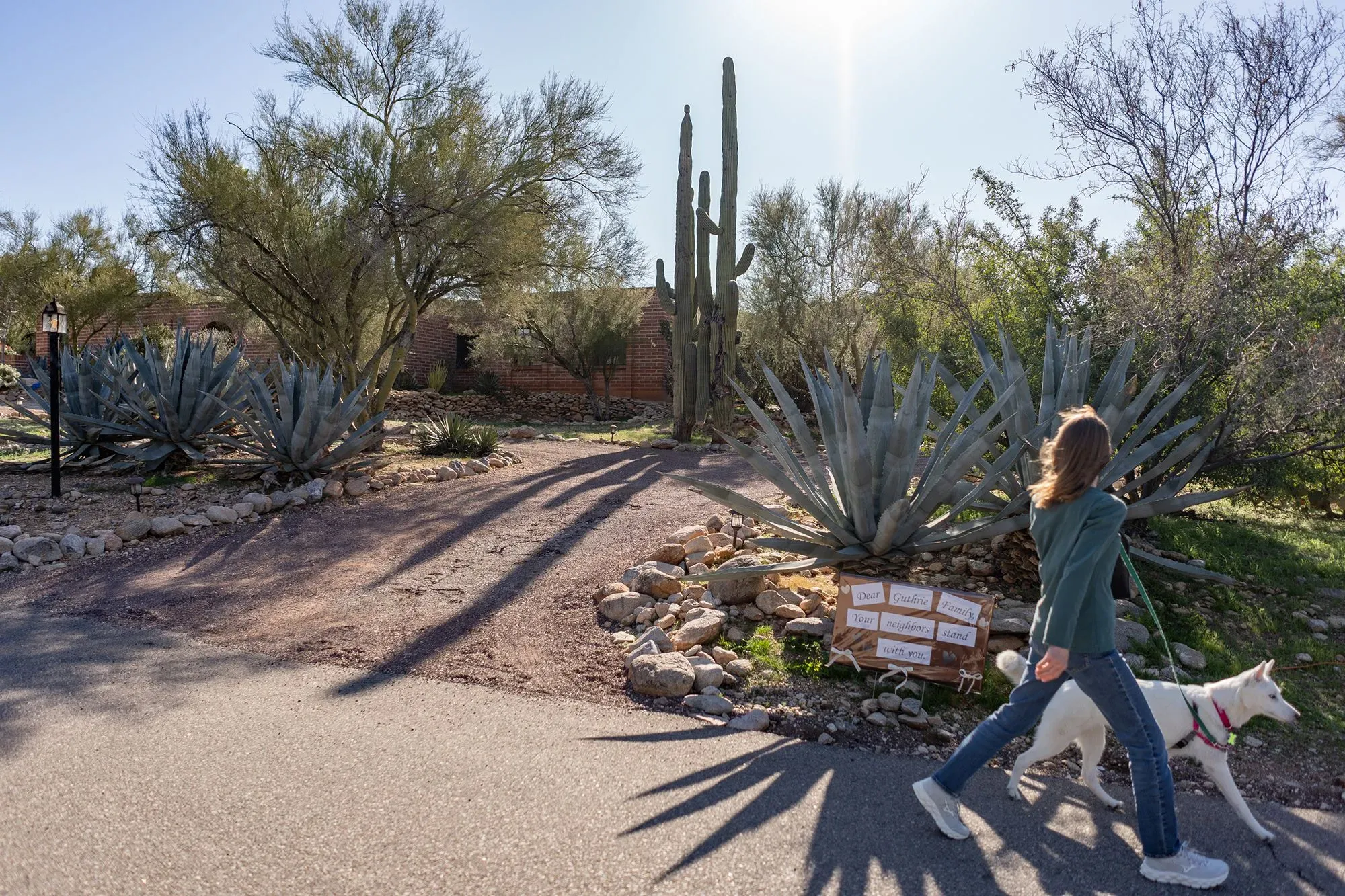 امرأة تسير مع كلبها في منطقة طبيعية، محاطة بأشجار وصبار، مع لافتات ترحب بالجيران. تعكس الصورة أجواء هادئة ومجتمعية.