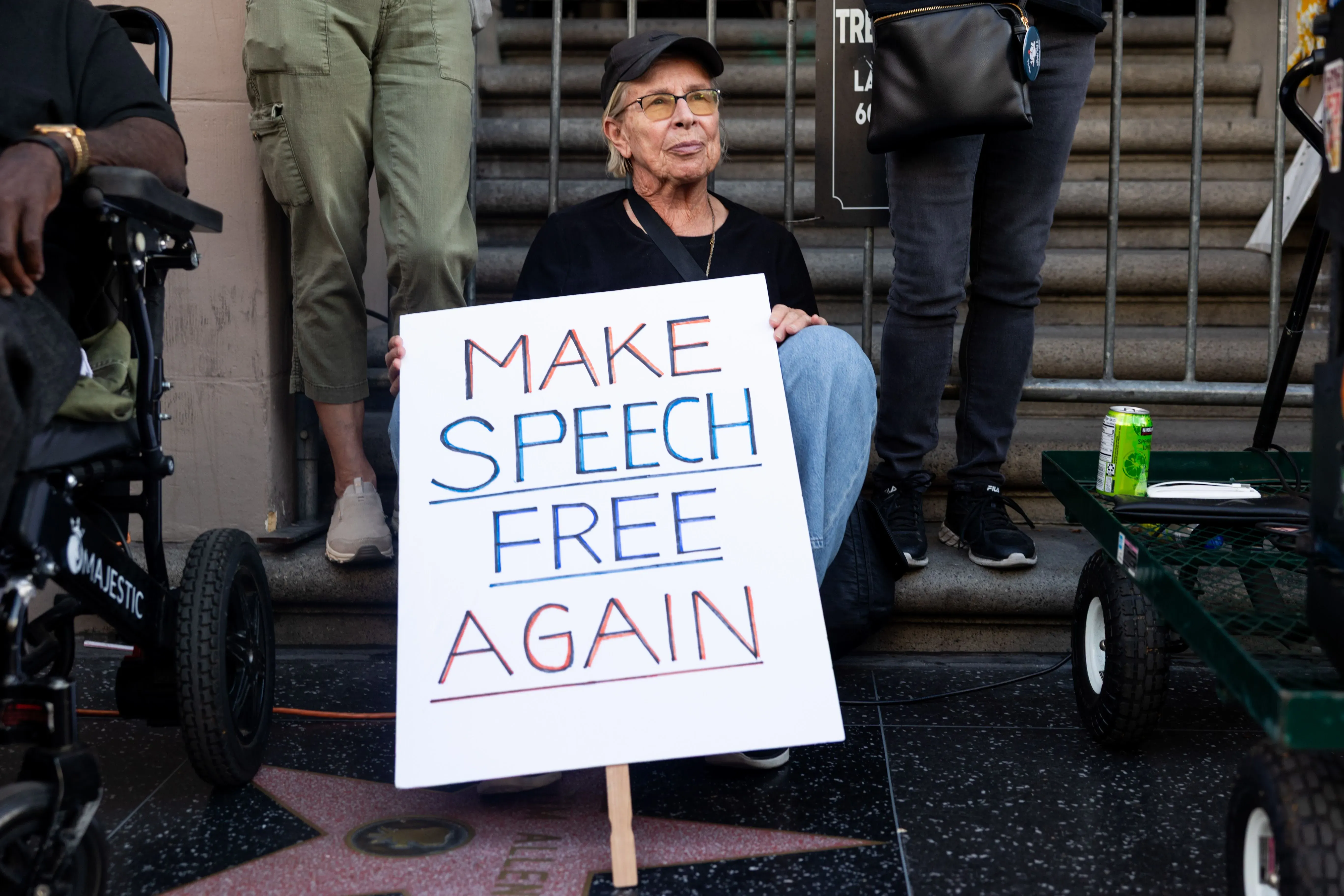 رجل مسن يجلس على درجات سلم حاملاً لافتة مكتوب عليها "اجعل الكلام حراً مرة أخرى"، بينما يقف آخرون بالقرب منه.