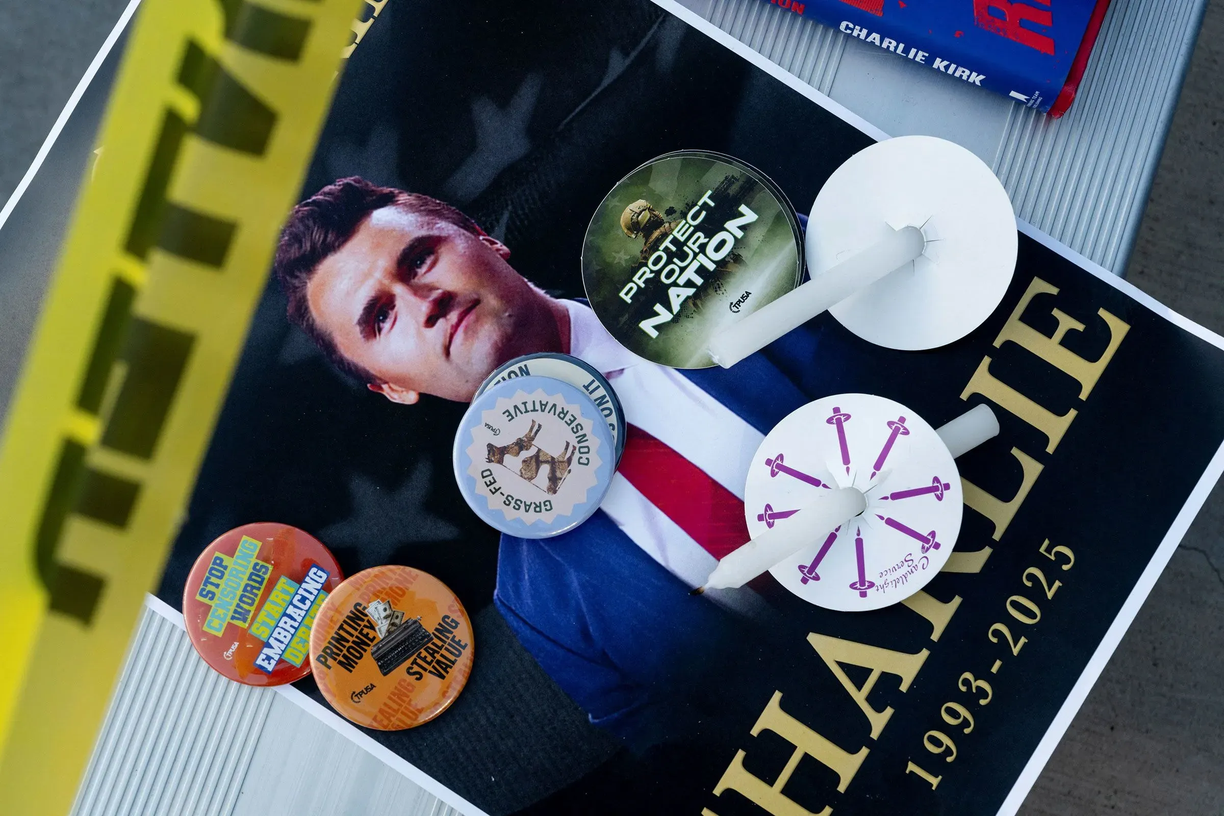 صورة تظهر ملصقًا يحمل صورة تشارلي كيرك، مع شارات تحمل شعارات سياسية، ومشغولات ضوئية، وخطوط صفراء تشير إلى موقع جريمة.