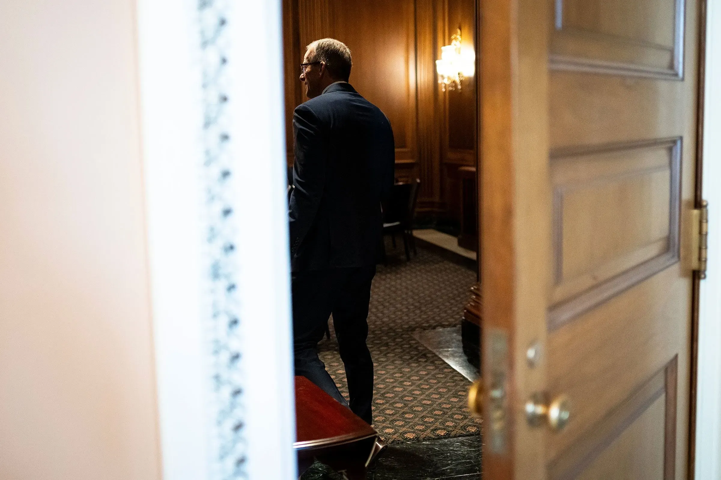 رجل يرتدي بدلة يغادر غرفة مغلقة في الكونغرس، مع خلفية من الأثاث الخشبي والإضاءة الدافئة، وسط أجواء من التوتر السياسي.