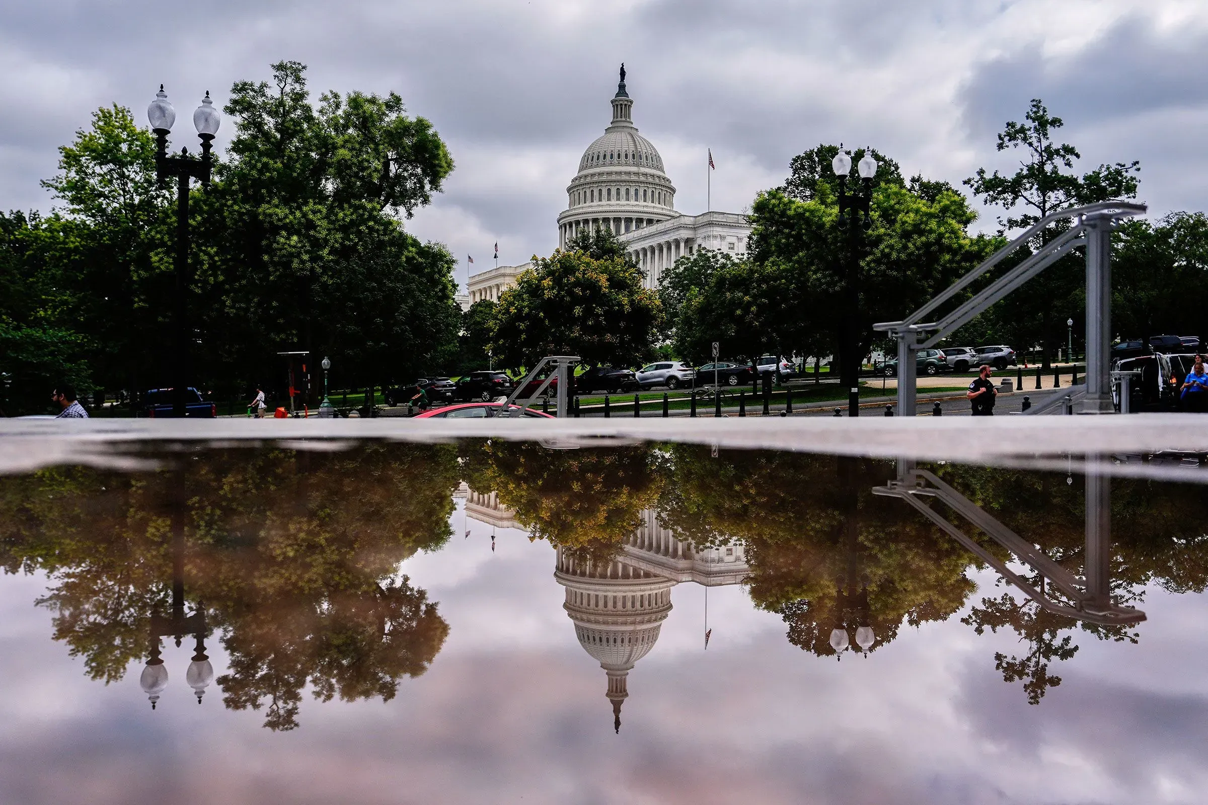 انعكاس مبنى الكابيتول الأمريكي في بركة ماء، مع سماء غائمة وأشجار محيطة، يرمز إلى التوترات السياسية المتزايدة في الكونغرس.