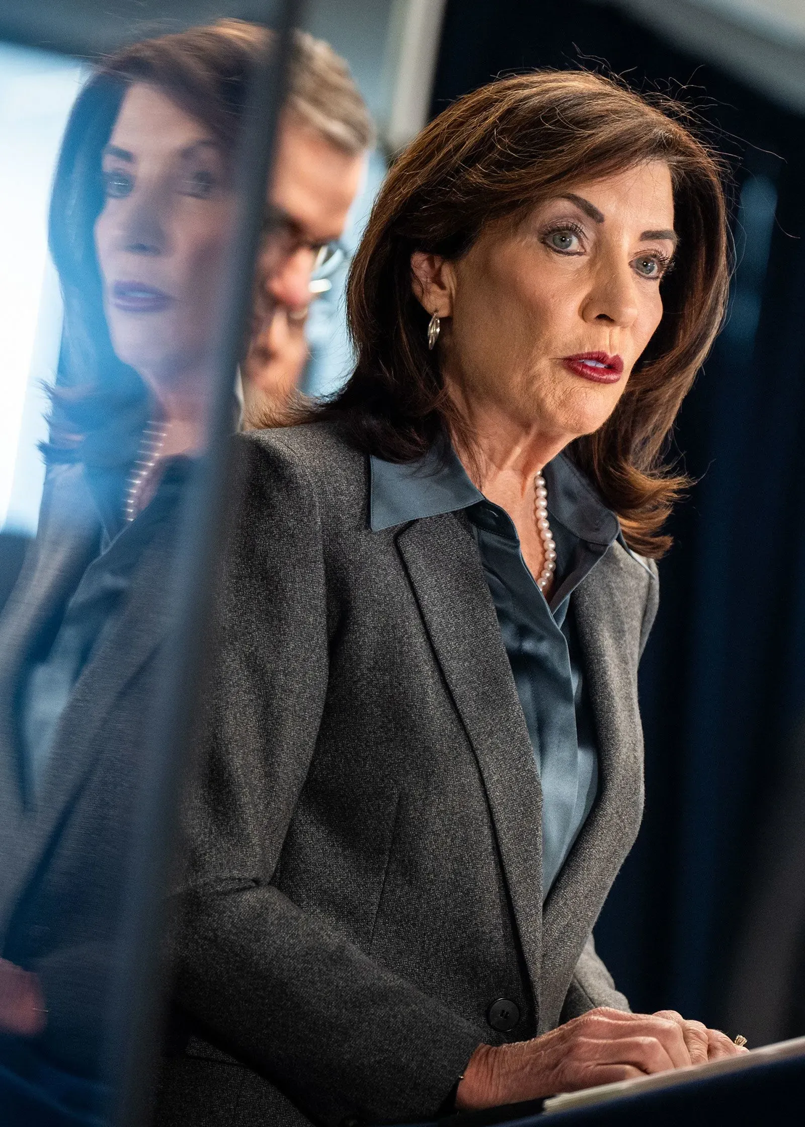 تظهر الصورة مسؤولة حكومية تتحدث خلال مؤتمر صحفي، مع التركيز على تعبير وجهها الجاد. تعكس الصورة التوترات حول سياسات الهجرة والتعاون مع السلطات الفيدرالية.