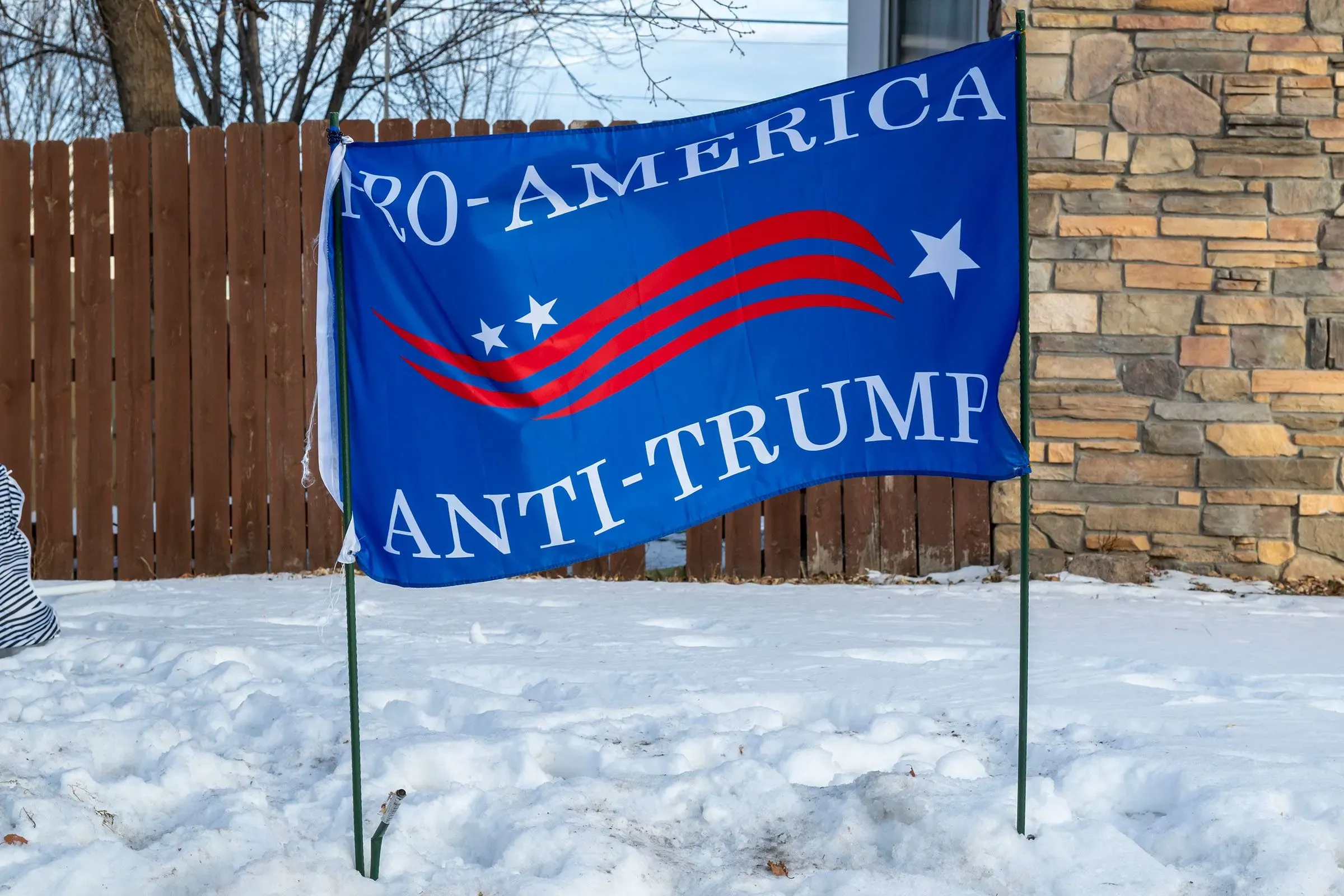 علم أزرق مكتوب عليه "مع أمريكا، ضد ترامب"، مثبت على عمود في منطقة مغطاة بالثلوج، مع خلفية من سياج خشبي.
