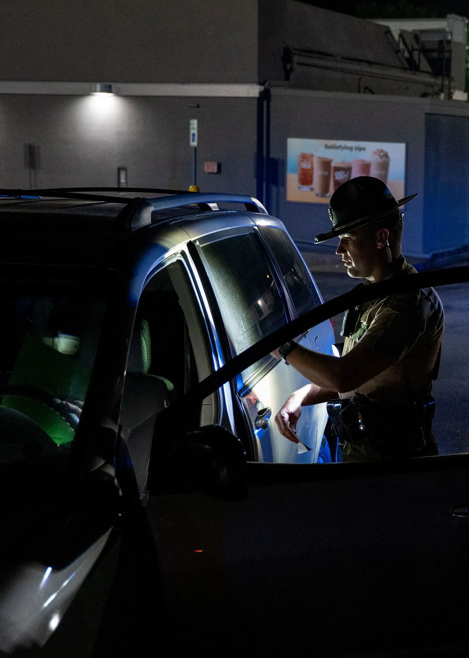 شرطي يقوم بتفتيش سيارة في الليل، مع خلفية مضاءة تشير إلى وجود لافتات إعلانية، مما يعكس جهود إنفاذ قوانين الهجرة.