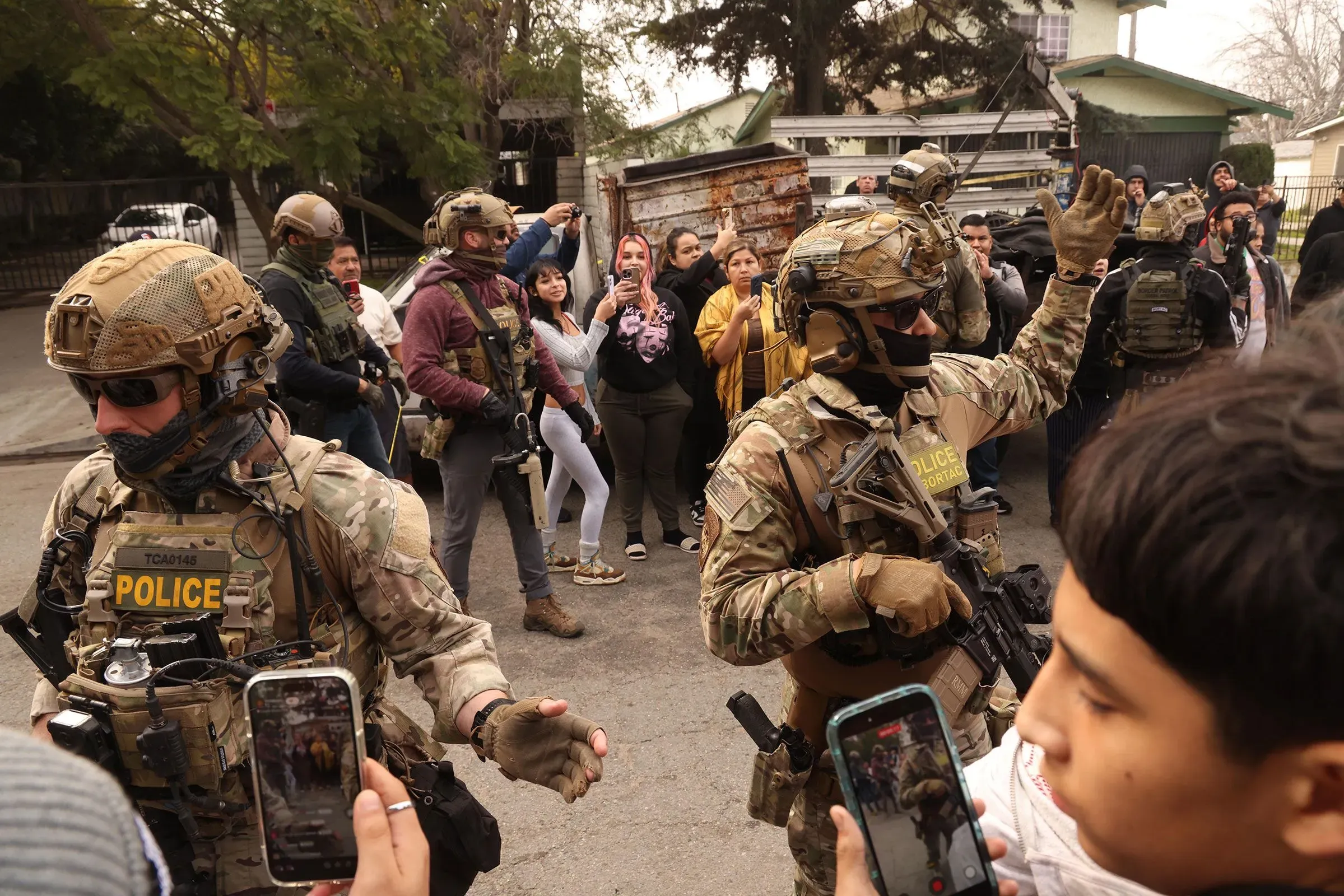 تظهر الصورة مجموعة من رجال الشرطة المدججين بالسلاح أثناء تنفيذ عملية في حي سكني، بينما يتجمع عدد من المواطنين لمشاهدة الحدث.