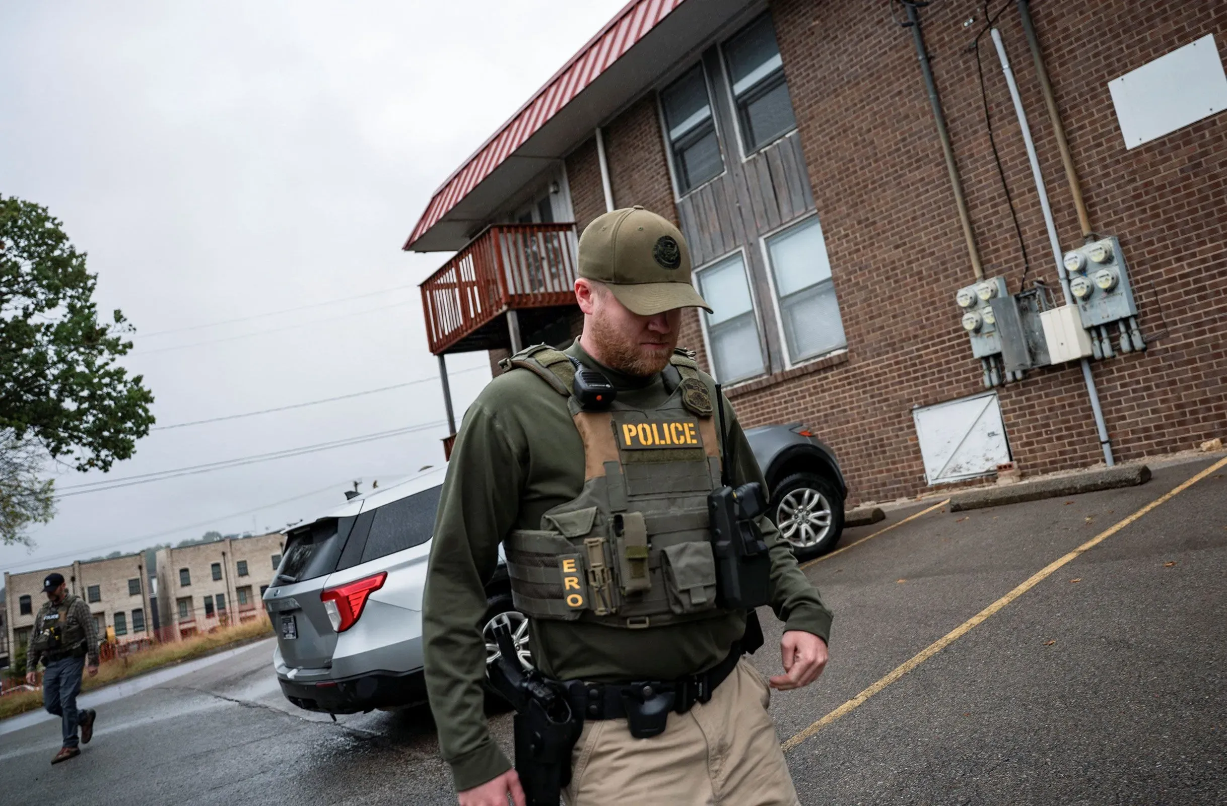 شرطي يرتدي سترة واقية وعلامة "شرطة" أثناء تفقده منطقة سكنية، في سياق تنفيذ قوانين الهجرة.