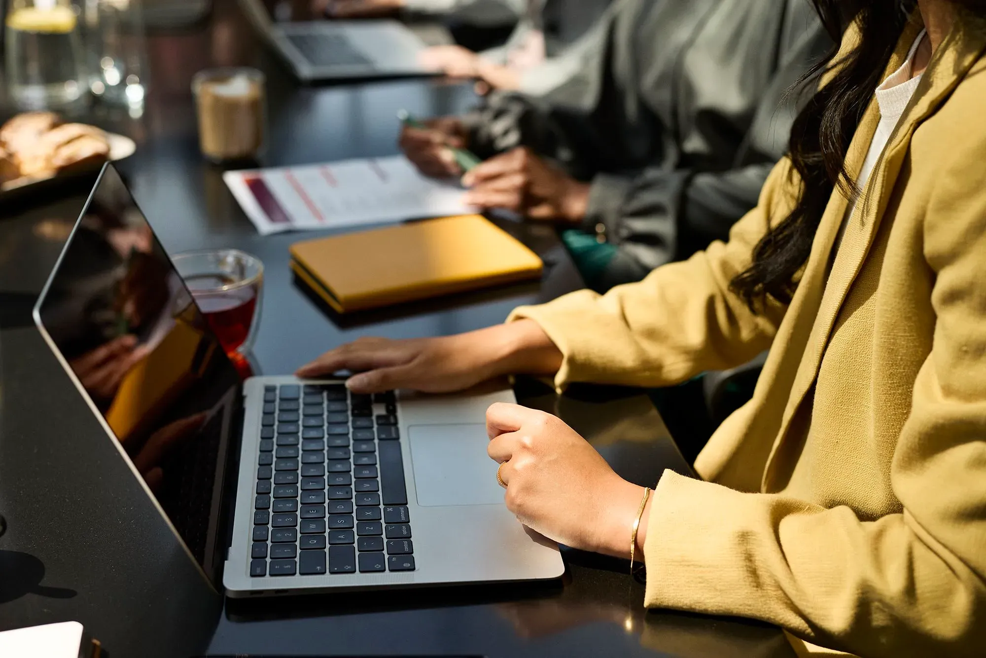 امرأة ترتدي سترة صفراء تعمل على حاسوب محمول في اجتماع، مع وجود مشروبات وملفات على الطاولة، مما يعكس بيئة عمل مهنية.