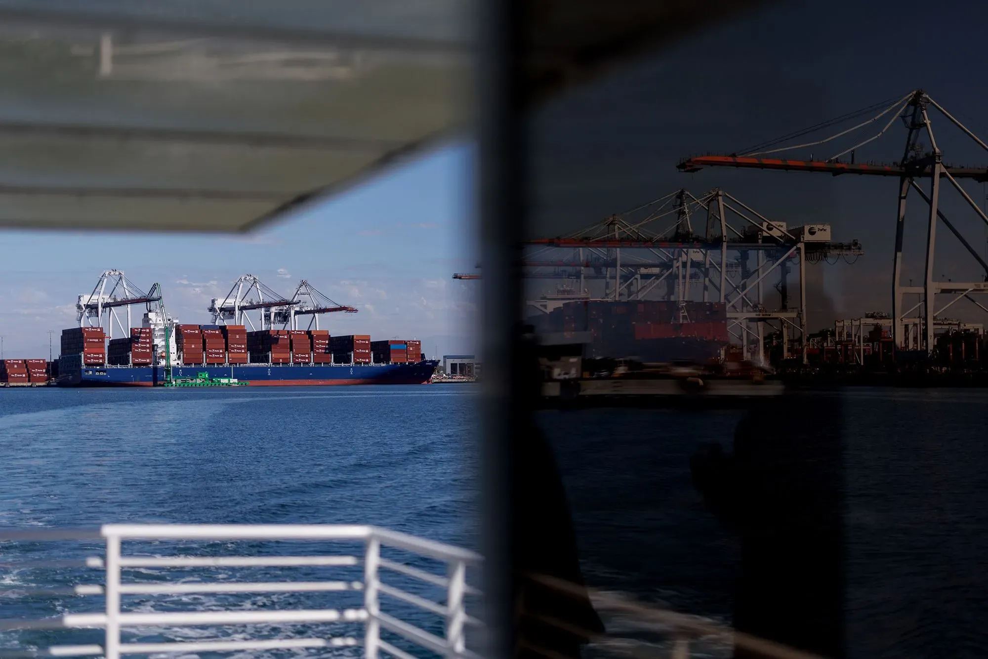 سفينة شحن محملة بالحاويات ترسو في ميناء، مع ظهور رافعات خلفية. تعكس الصورة التوترات التجارية والرسوم الجمركية في ظل إدارة ترامب.