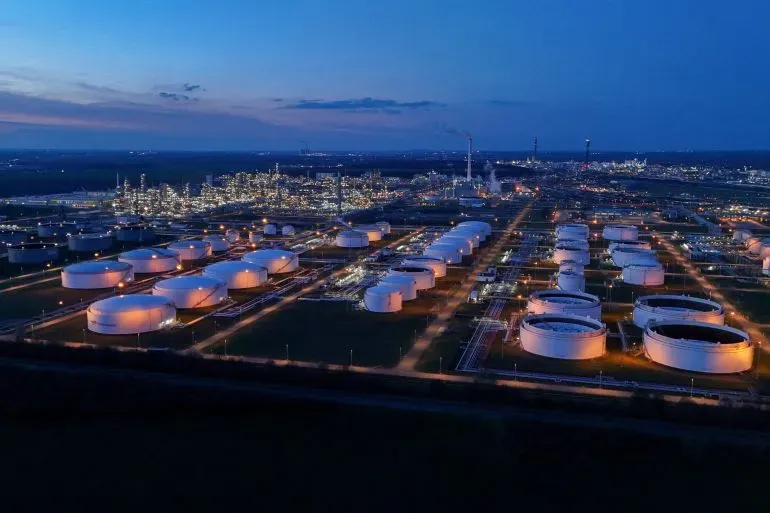 منشأة تخزين نفطية كبيرة مضاءة في الليل، مع وجود صهاريج ضخمة ومصانع في الخلفية، تعكس تأثير ارتفاع أسعار النفط على السوق العالمية.