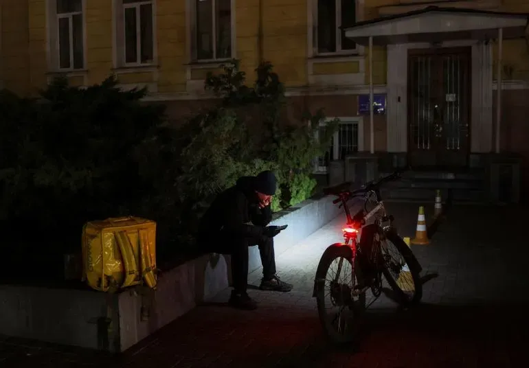 رجل يرتدي قبعة سوداء يجلس في الشارع ليلاً، يستخدم هاتفه المحمول بجوار دراجة، مع وجود حقيبة صفراء مضاءة في الخلفية.