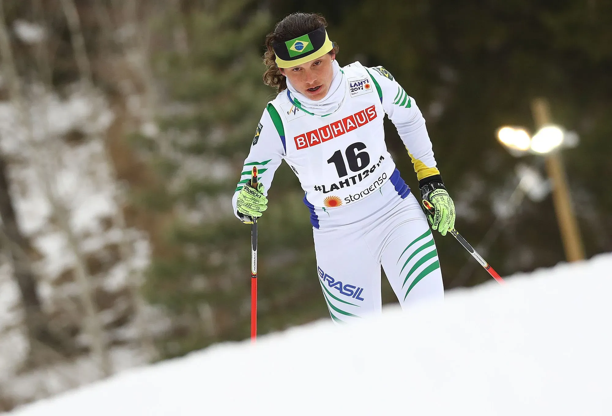 برازيليّة تتزلج على الثلج، ترتدي زيًّا رياضيًا يحمل الرقم 16، تتسلق منحدرًا ثلجيًا في بطولة التزلج. تعكس الصورة عزيمتها في تحقيق حلمها الأولمبي.