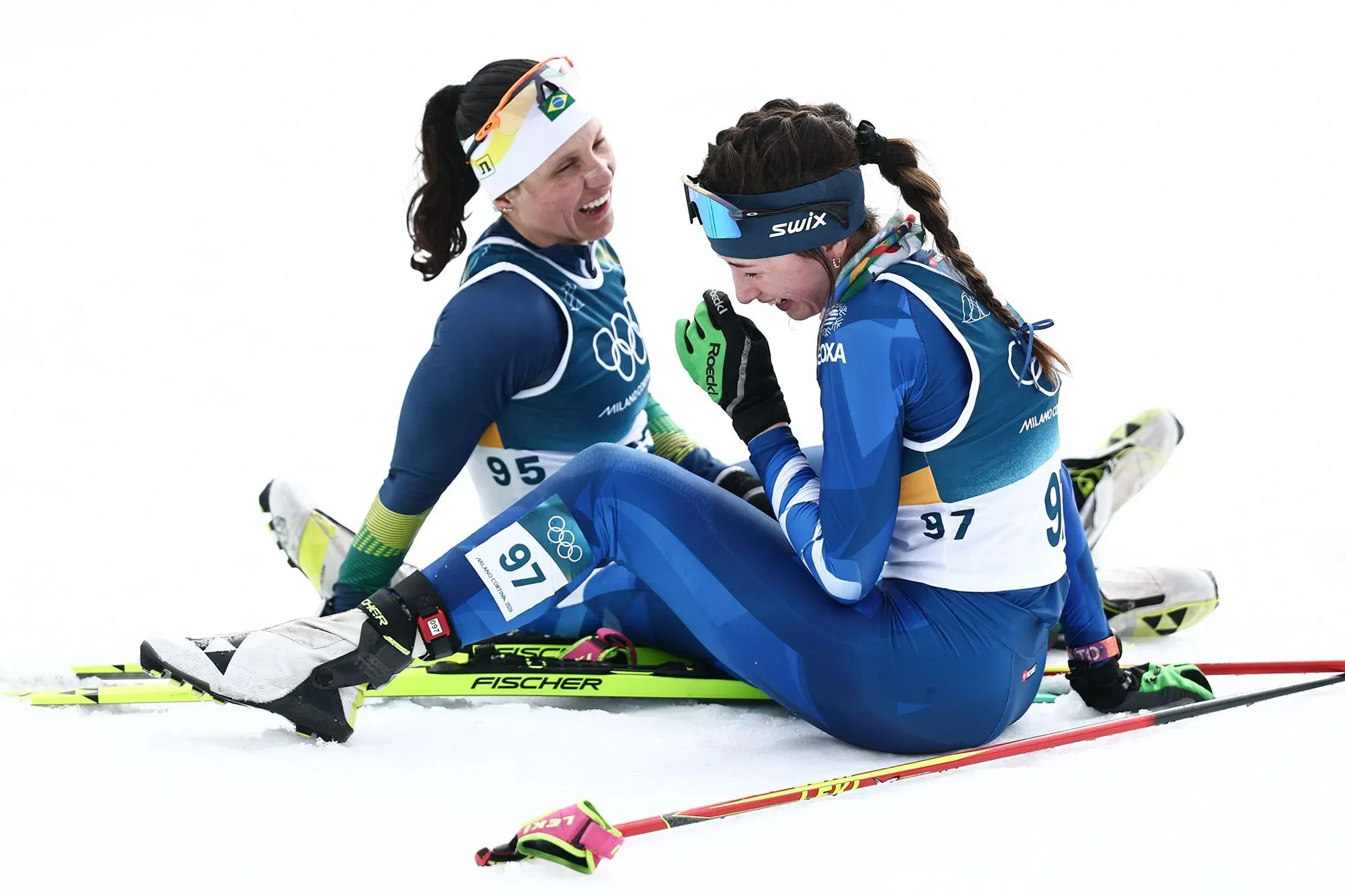 برازيليتان تتبادلان الضحك بعد المنافسة في التزلج الريفي على الثلج، مع ارتداء ملابس رياضية زرقاء، تعبيرات الفرح تبرز روح الأولمبياد.