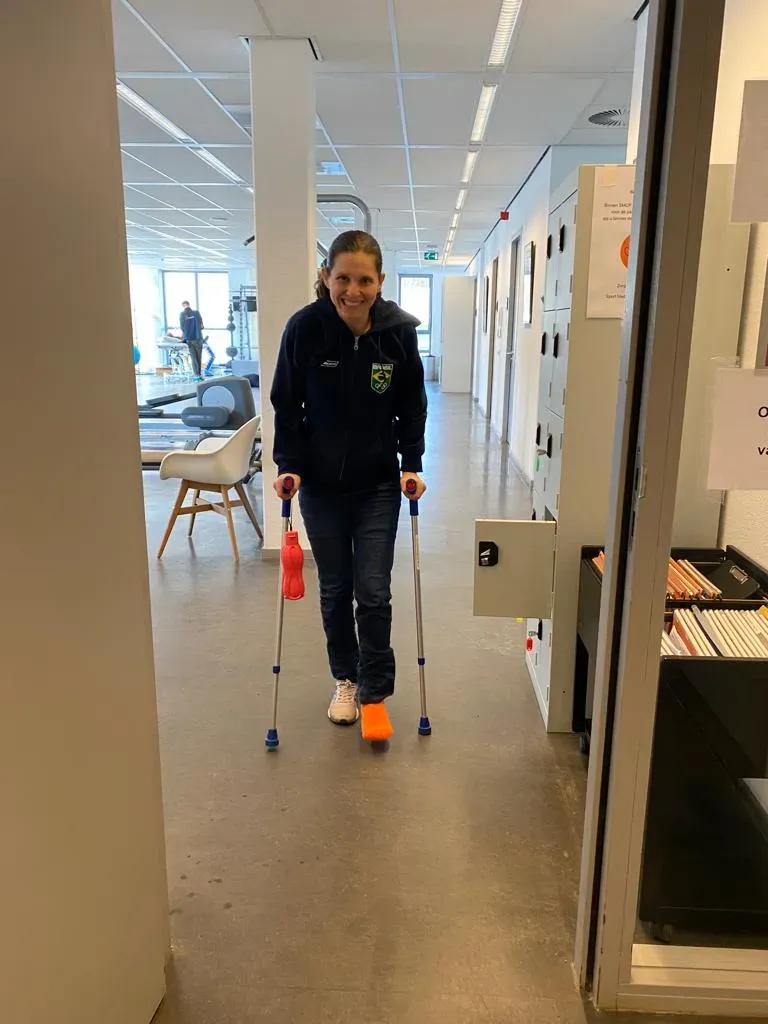 امرأة تستخدم عكازين في ممر داخلي، مبتسمة، ترتدي سترة رياضية تحمل شعار البرازيل، تعكس روح العزيمة والتحدي في مسيرتها الرياضية.