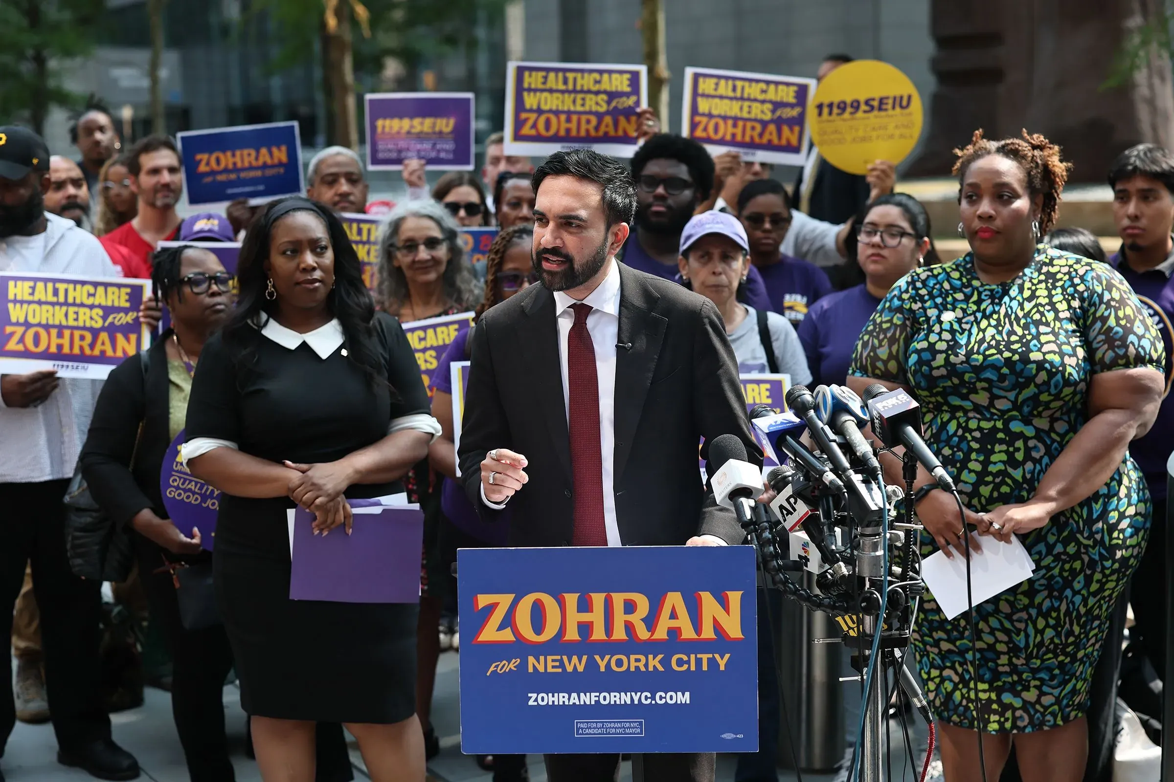 مؤتمر صحفي لزهران ممداني في مانهاتن، حيث يتحدث عن مستقبل المدينة محاطًا بأعضاء من نقابة عمال الرعاية الصحية، مع لافتات تدعمه.
