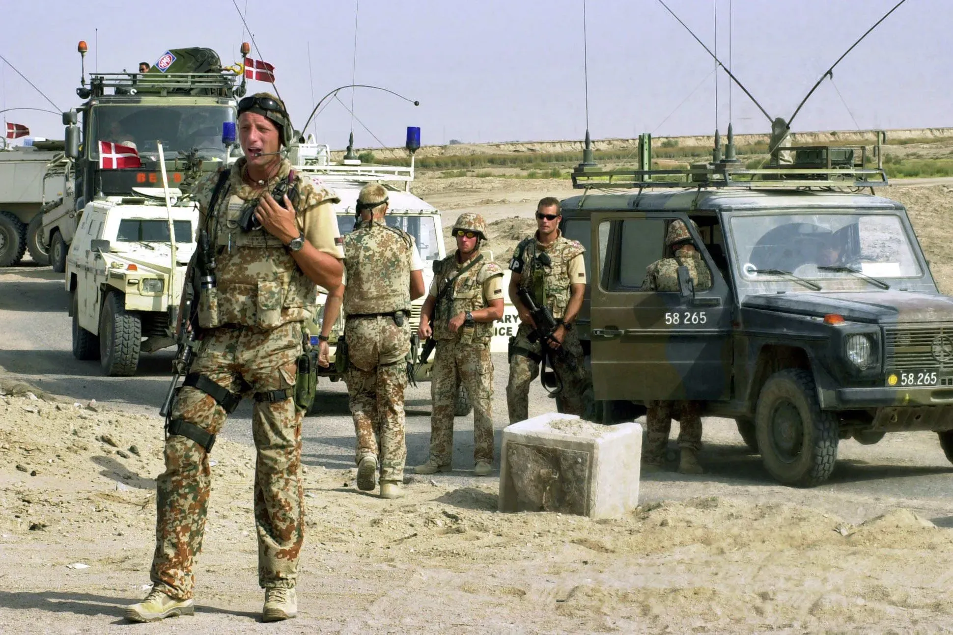 جنود دنماركيون في مهمة عسكرية، يرتدون زيهم الرسمي، يتجمعون بالقرب من مركباتهم في منطقة صحراوية، مع أعلام دنماركية ترفرف.