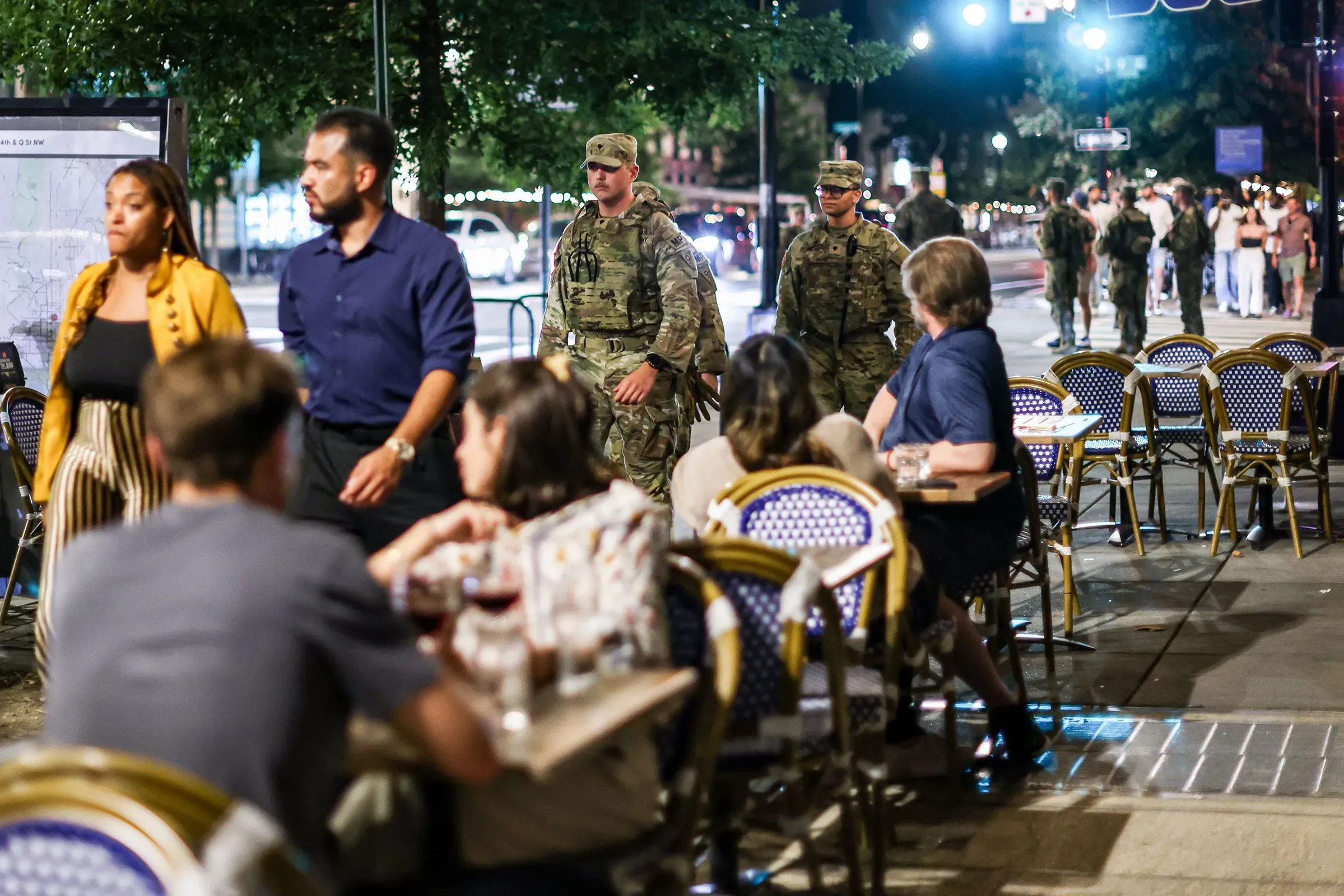 تظهر الصورة جنودًا يرتدون زيًا عسكريًا يتجولون في شوارع واشنطن العاصمة، بينما يتناول بعض الأشخاص الطعام في المطاعم المجاورة.