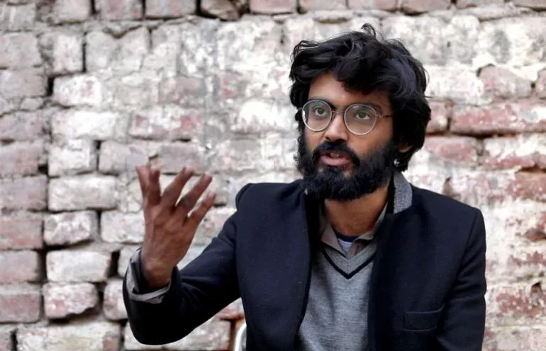 ناشط يتحدث بجدية، يرتدي نظارات، وخلفه جدار من الطوب. الصورة تعكس قضايا العدالة الاجتماعية وحقوق المسلمين في الهند.